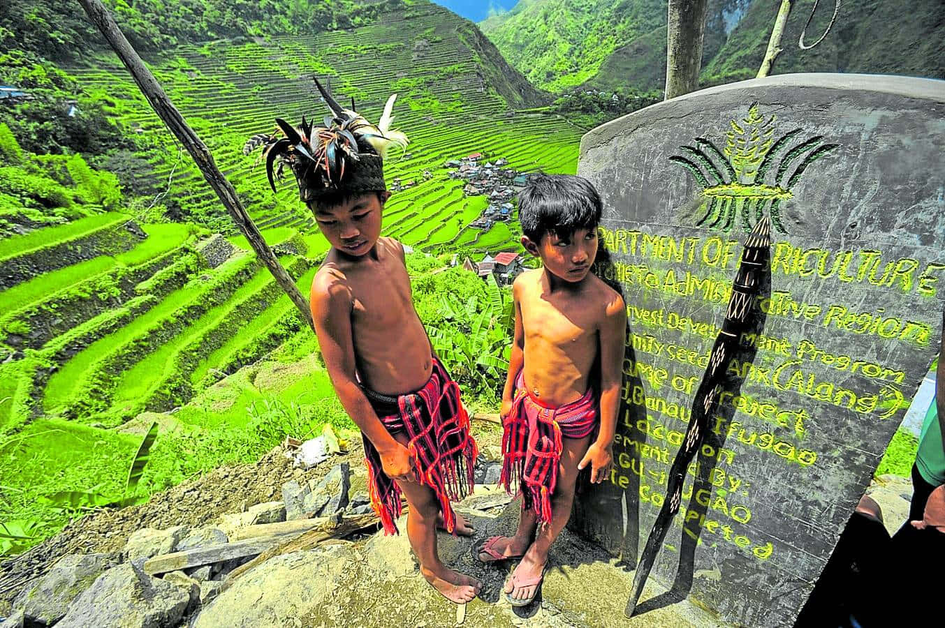 兩名身穿傳統服飾的年輕原住民男孩站在標誌附近，俯瞰梯田。