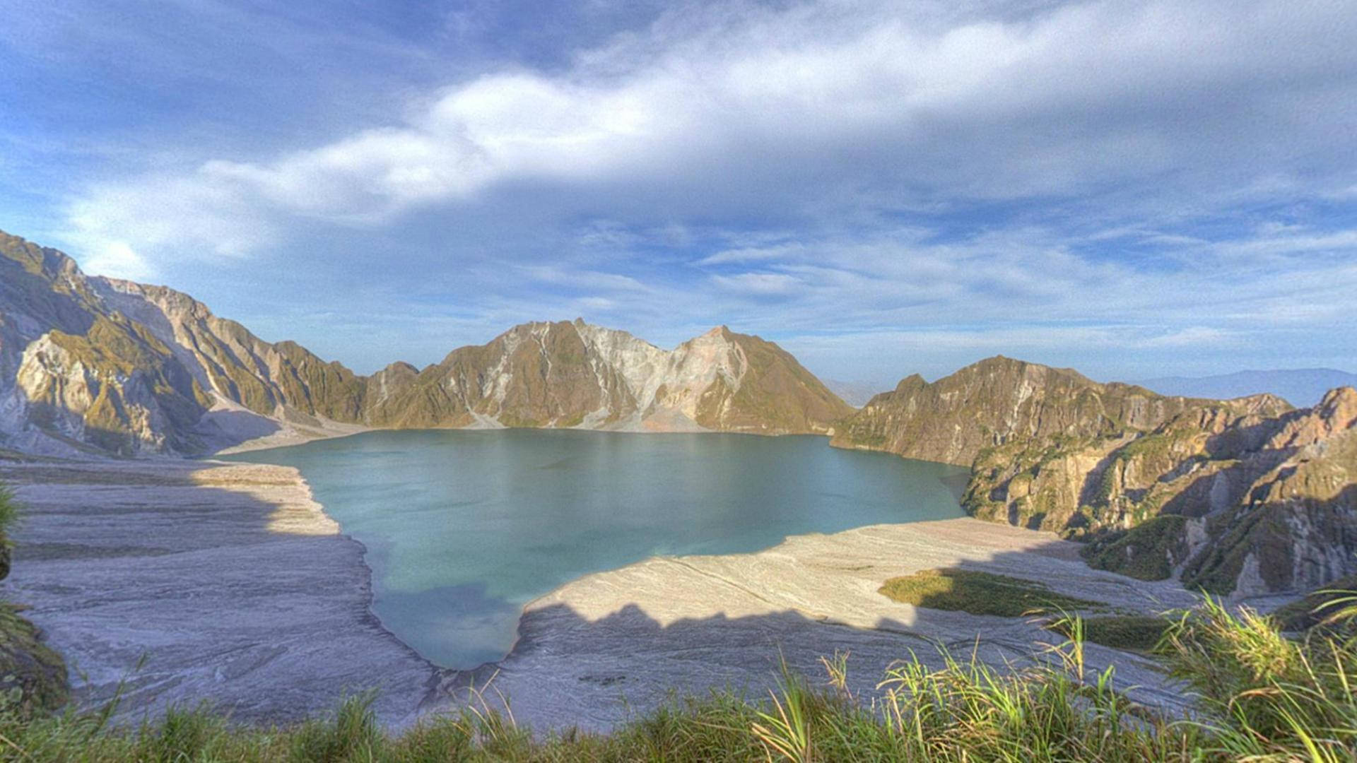 菲律賓皮納圖博湖 壁紙 — 一個寧靜的火山口湖，被崎嶇的山脈環繞，在明亮的藍天和零星雲朵下。