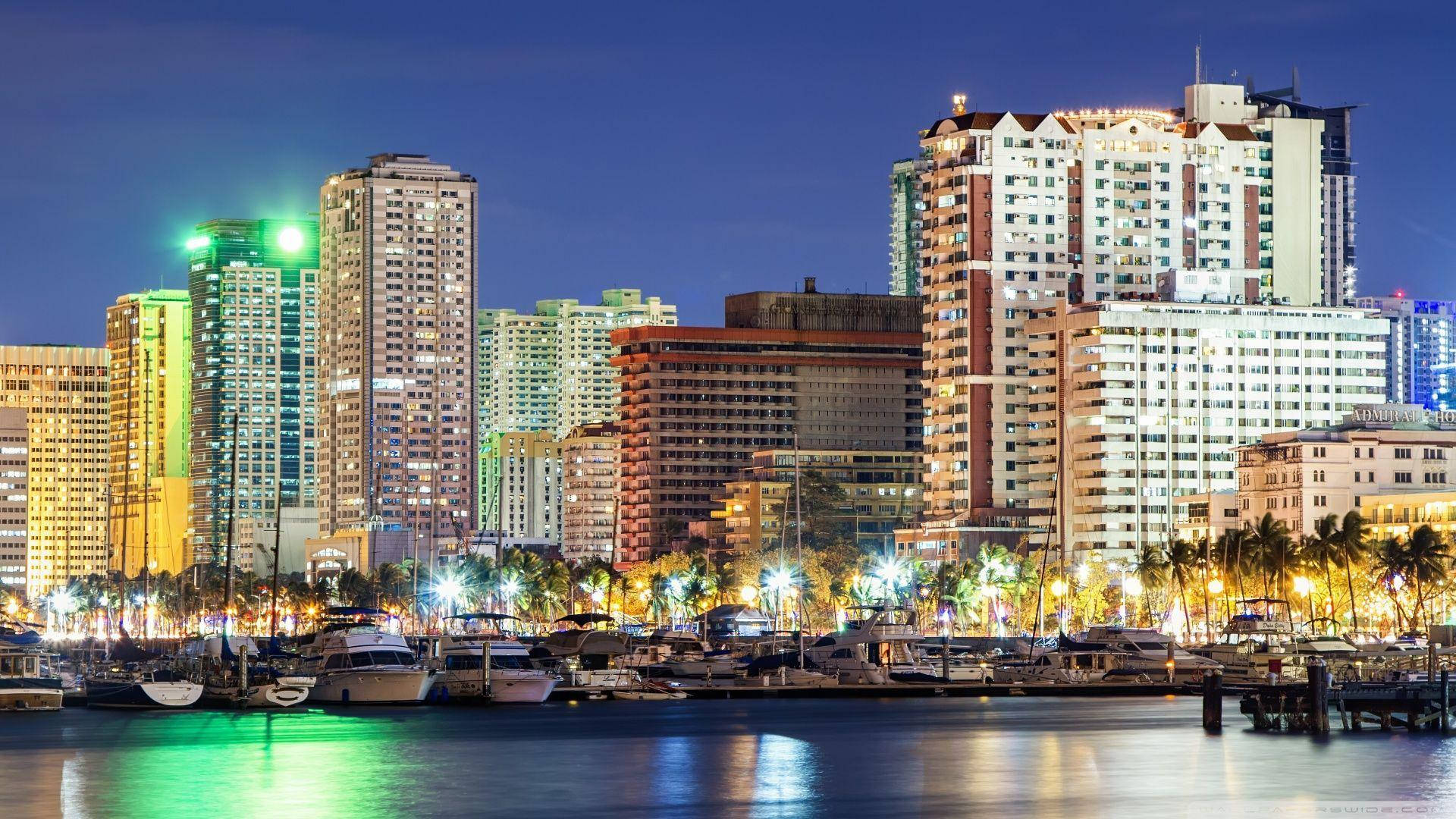 菲律賓羅克薩斯大道夜景 壁紙 — 充滿活力的夜景城市景觀，燈火通明的摩天大樓倒映在平靜的水面上，旁邊是停滿船隻的碼頭。