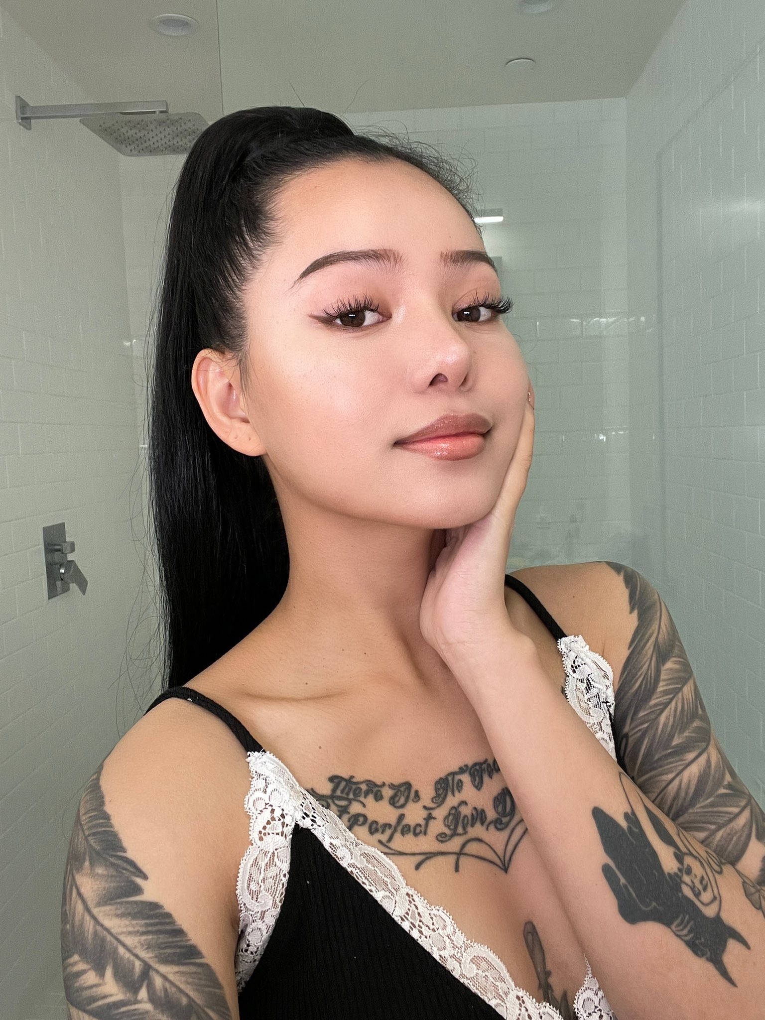 菲律賓裔美國人Bella Poarch 壁紙 — 一位長黑髮及有紋身的女性在白色瓷磚浴室中穿著黑色蕾絲上衣擺姿勢。