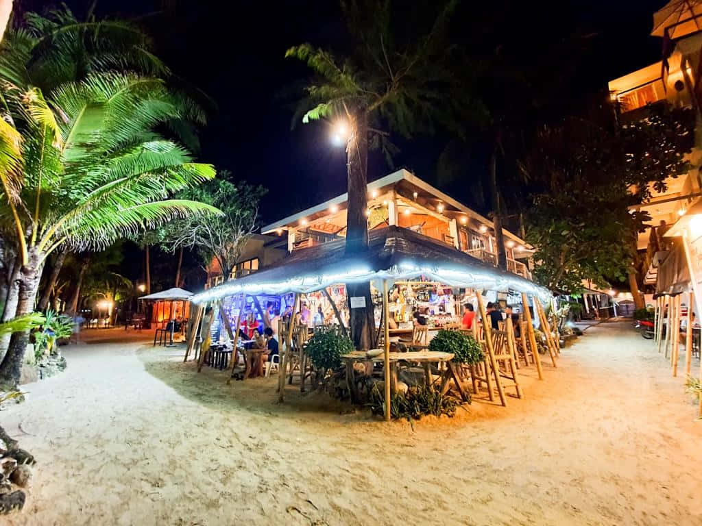 菲律賓長灘島夜間海灘 壁紙 — 海邊露天酒吧在夜晚棕櫚樹下溫暖地發光，客人在沙灘上享用飲品。