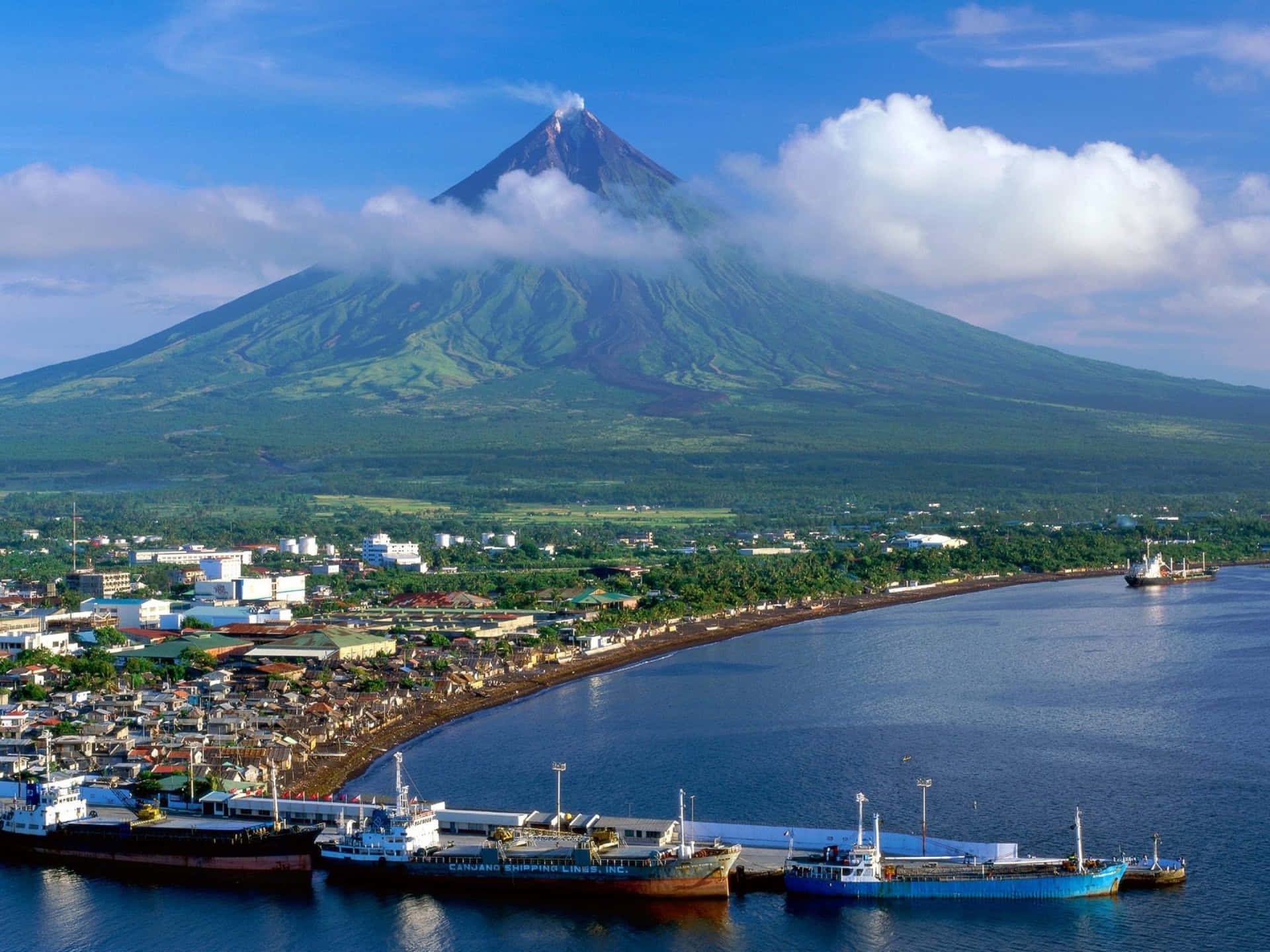 沿海小鎮，船隻停泊在藍天下的大型綠色火山附近。