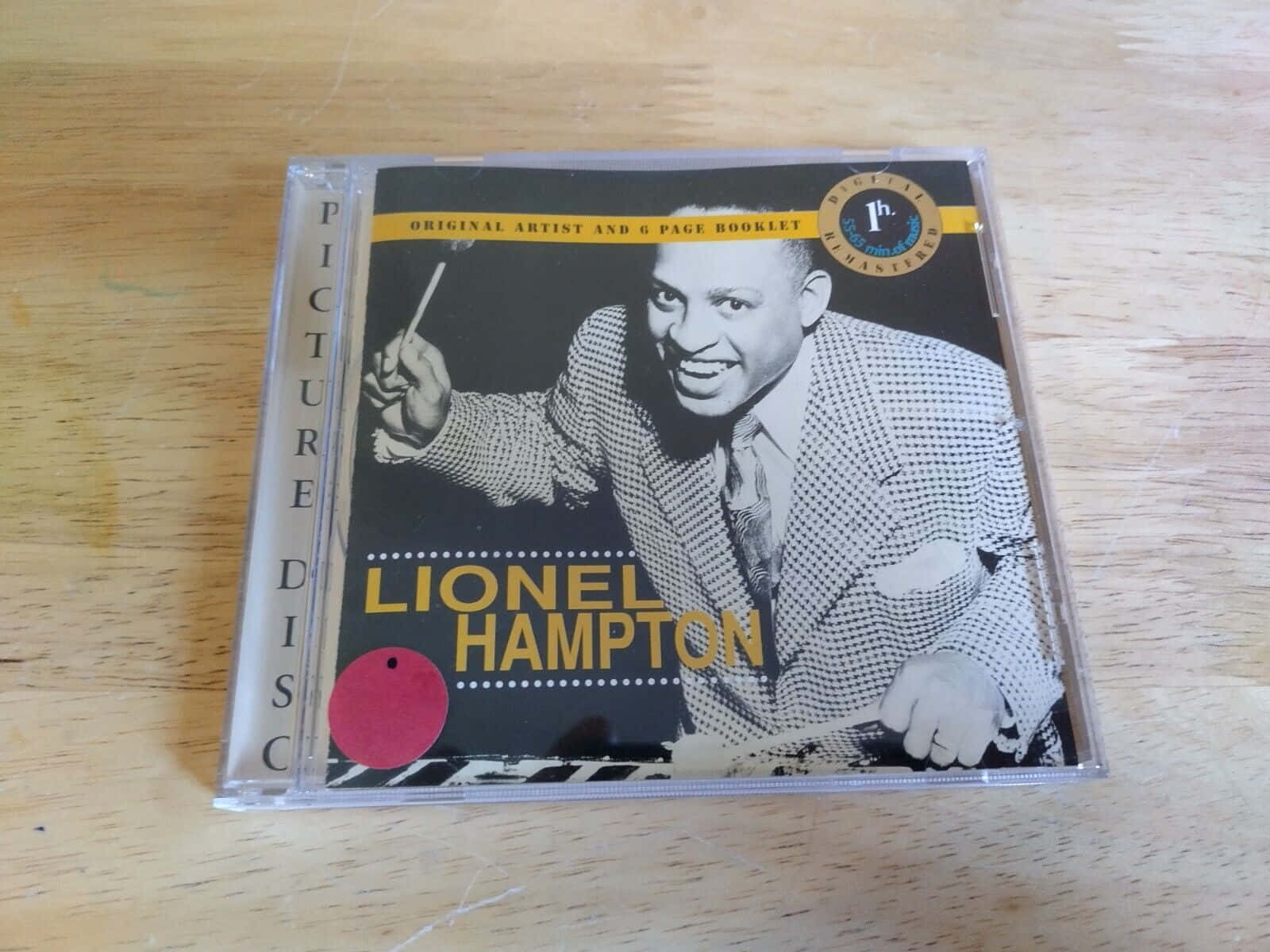 萊昂內爾·漢普頓《熱力鼓棒》專輯封面 壁紙 — 一張Lionel Hampton微笑握住鼓棒的復古圖案CD放在淺色木質表面上。