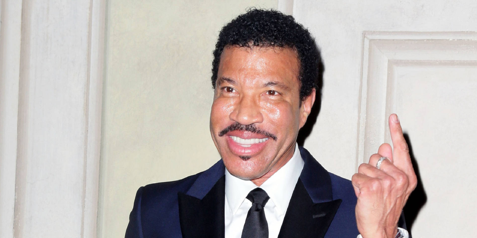 Lionel Richie 身穿深色西裝，在室內微笑並向上指。