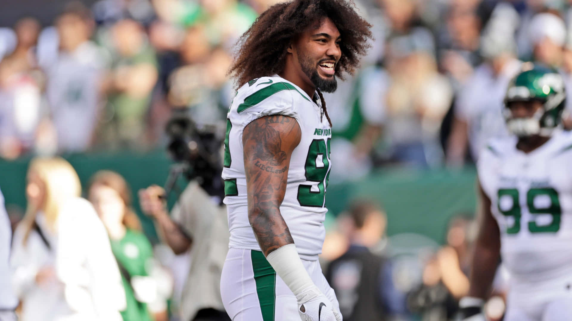萊昂納德·威廉斯紐約巨人隊足球 壁紙 — 一位微笑的NFL球員，留著捲髮和紋身，在陽光明媚的足球場上穿著綠色和白色的裝備慶祝。