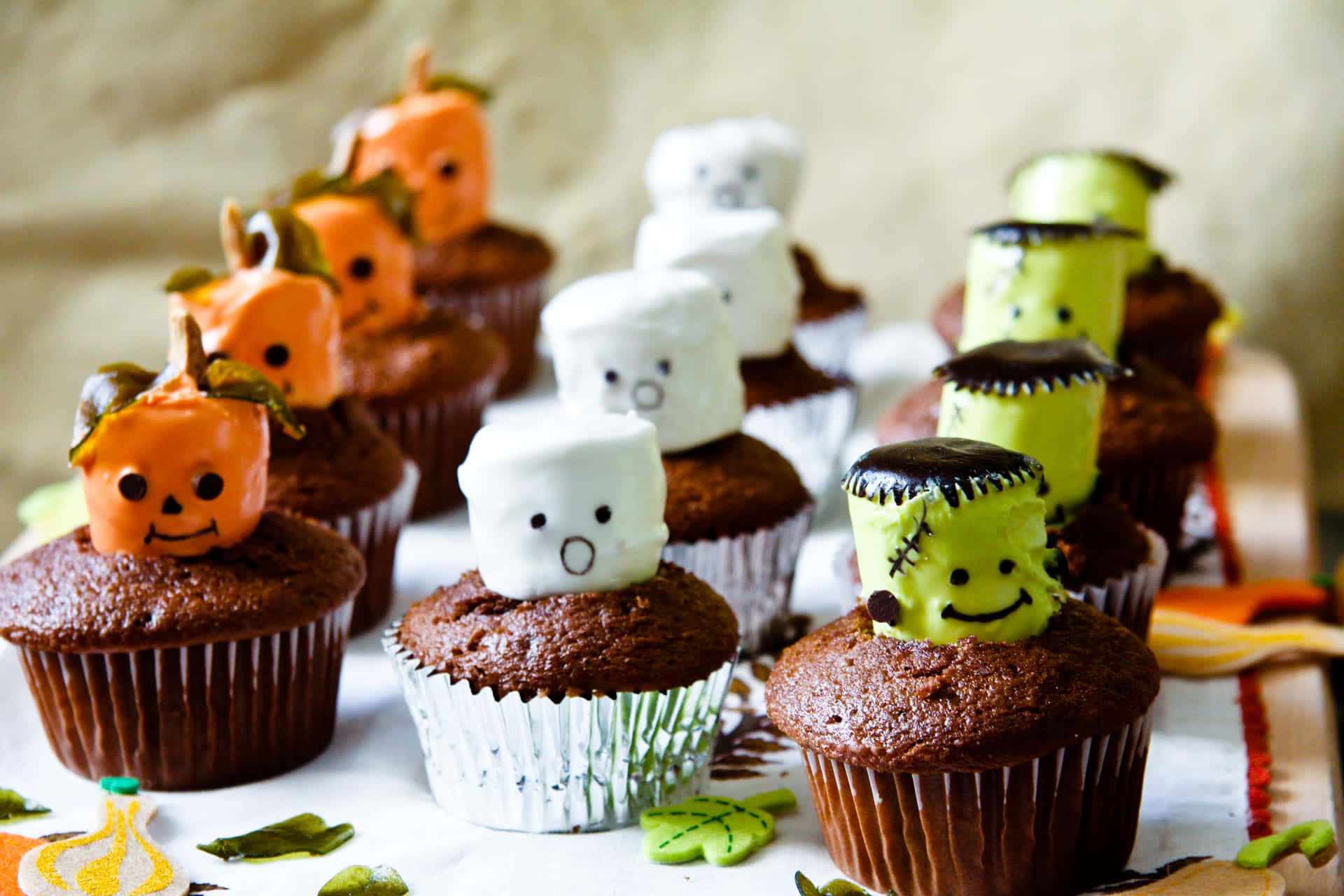 Halloween Cupcakes 桌布 壁紙 — 萬聖節主題紙杯蛋糕，上面放有橙色南瓜棉花糖、白色鬼魂棉花糖和綠色科學怪人棉花糖。