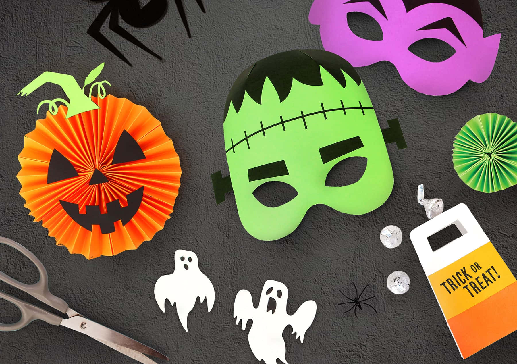 Halloween Crafts 桌布 壁紙 — 萬聖節手工藝品的平鋪擺放，包括綠色科學怪人面具、橙色紙南瓜、白色鬼魂和一個不給糖就搗蛋的袋子，放在深色紋理表面上。