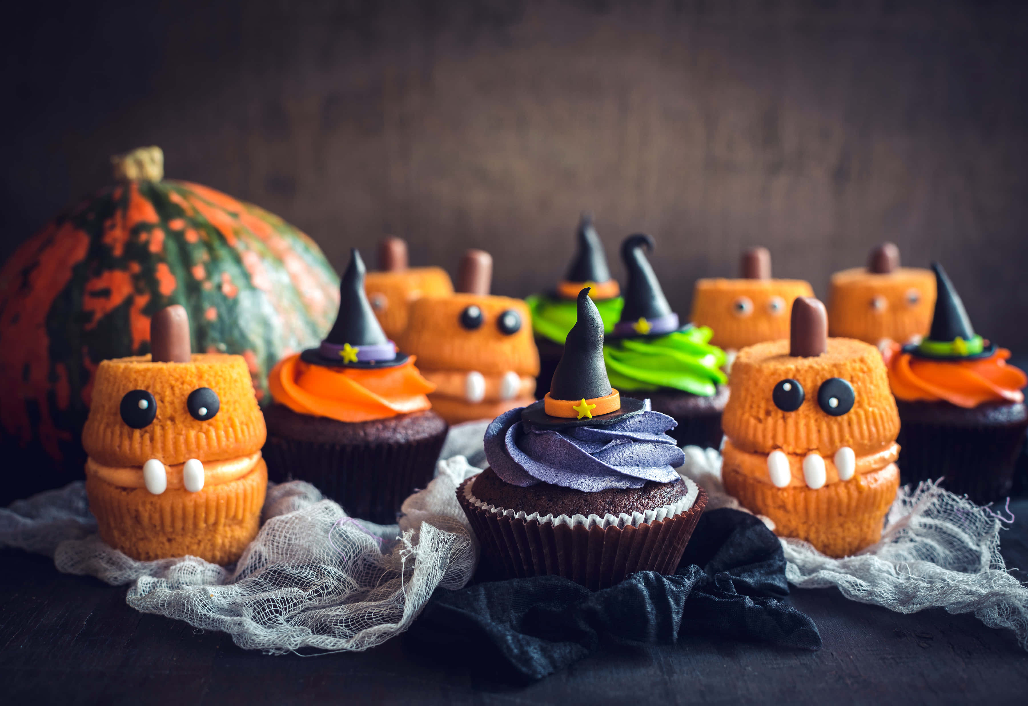Halloween Cupcakes 桌布 壁紙 — 萬聖節杯子蛋糕裝飾成南瓜和女巫，配上橙色糖霜和黑色帽子，放在黑暗的桌子上，旁邊有一個真正的南瓜。