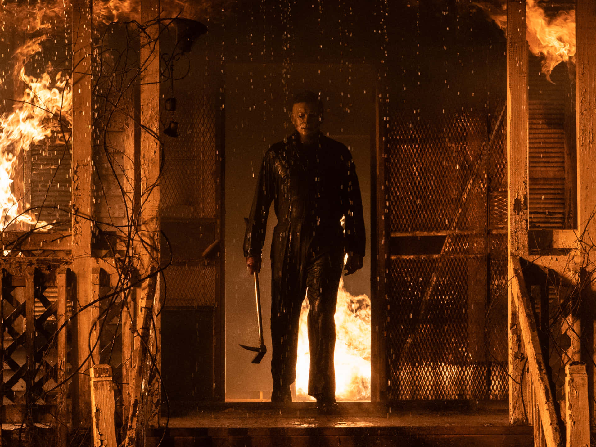 萬聖節邁克爾·邁爾斯在燃燒的房屋前 壁紙 — Michael Myers 企喺被雨水淋濕嘅門口，身後嘅木造門廊喺黑暗天空下被火焰吞噬。