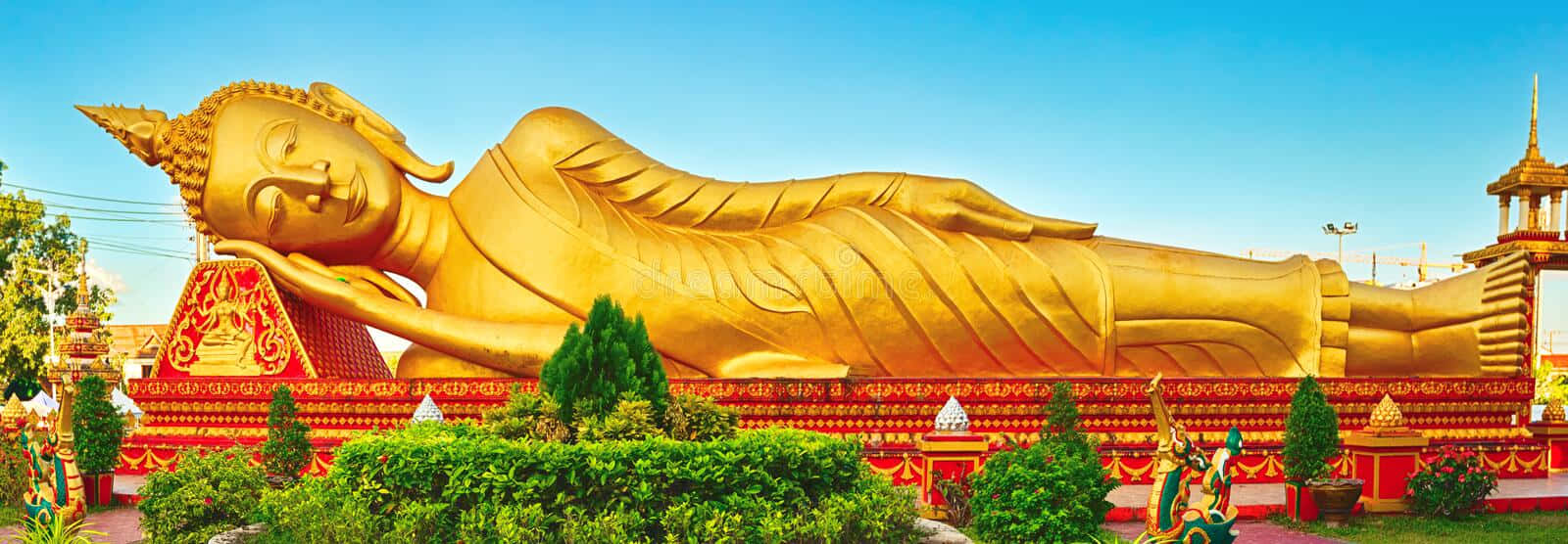 萬象的金色佛塔 壁紙 — 一尊巨大的金色臥佛在晴朗的藍天下安詳地躺著，周圍有鮮紅色的寺廟建築和茂密的綠色植物。