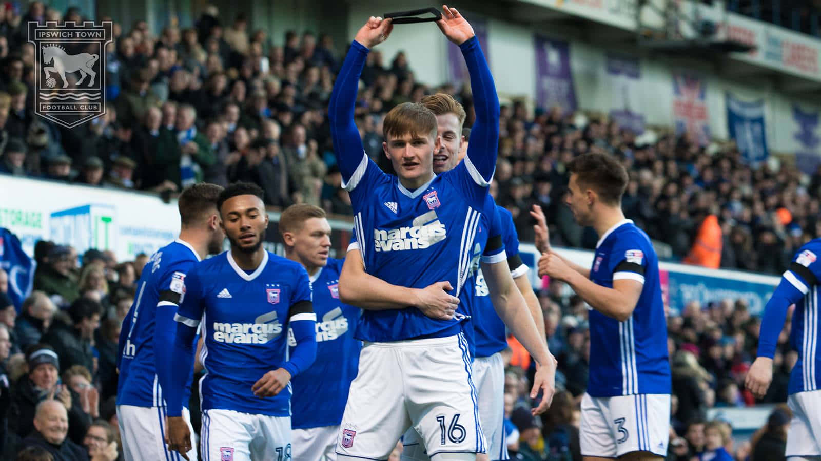 Ipswich Town 桌布 壁紙 — 葉士域治足球員在擠滿觀眾的球場前慶祝入球，身穿藍色球衣。