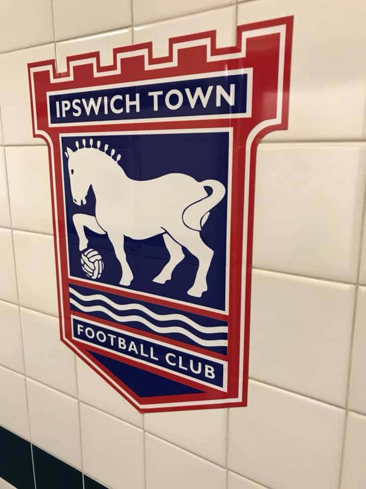 Ipswich Town 桌布 壁紙 — 葉士域治足球會徽章，上有白馬和足球，藍色背景，安裝在白色瓷磚牆上。