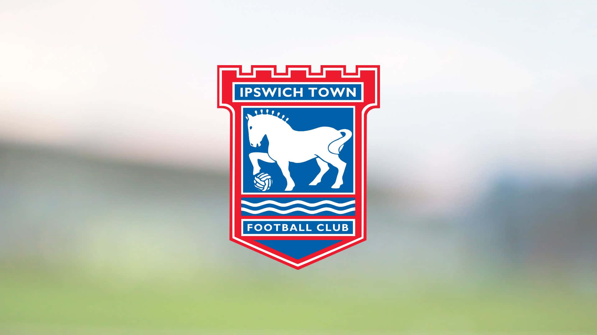 Ipswich Town 桌布 壁紙 — 葉士域治足球會徽章，藍色盾牌上有白馬和足球，背景為模糊的綠色球場。