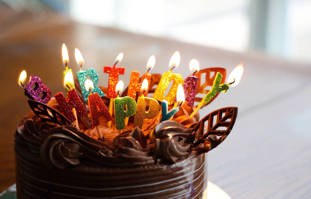 葉子設計生日蛋糕與蠟燭 壁紙 — 朱古力生日蛋糕特寫，上面有色彩繽紛閃爍的蠟燭拼出「HAPPY BIRTHDAY」字樣，並燃點著火焰。