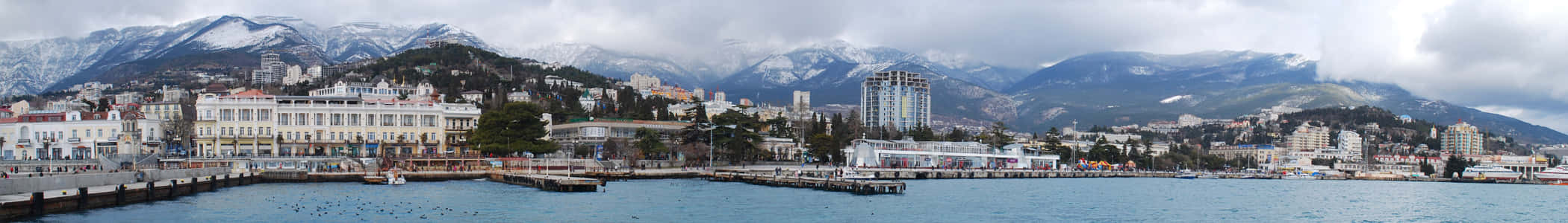 葉爾加瓦全景 壁紙 — 海岸城市的全景視圖，背景是白雪覆蓋的山脈，前景是平靜的水面，頭頂是陰天。