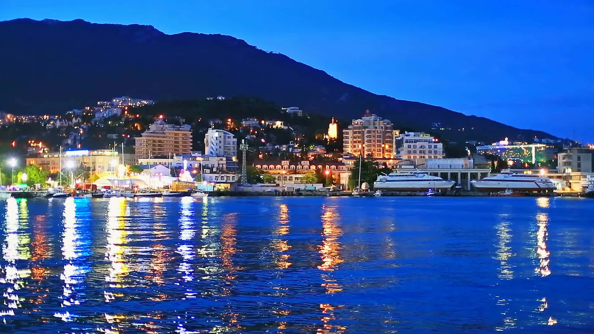 葉爾加瓦夜景全景 壁紙 — 暮色中的沿海城市，燈光閃爍，倒映在平靜的水面上，以黑暗的山脈為背景。