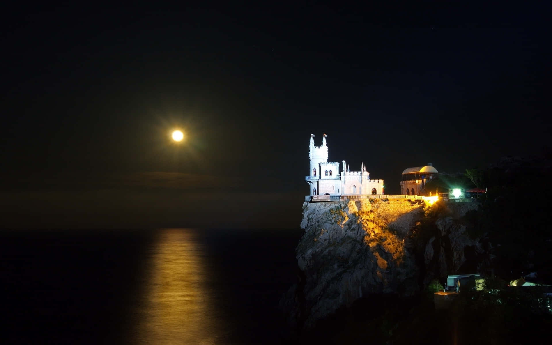 葉爾加瓦燕子城堡夜景 壁紙 — 一座宏偉的燈光城堡坐落在懸崖上，夜晚明亮的滿月在下方水面上投下金色的光芒。