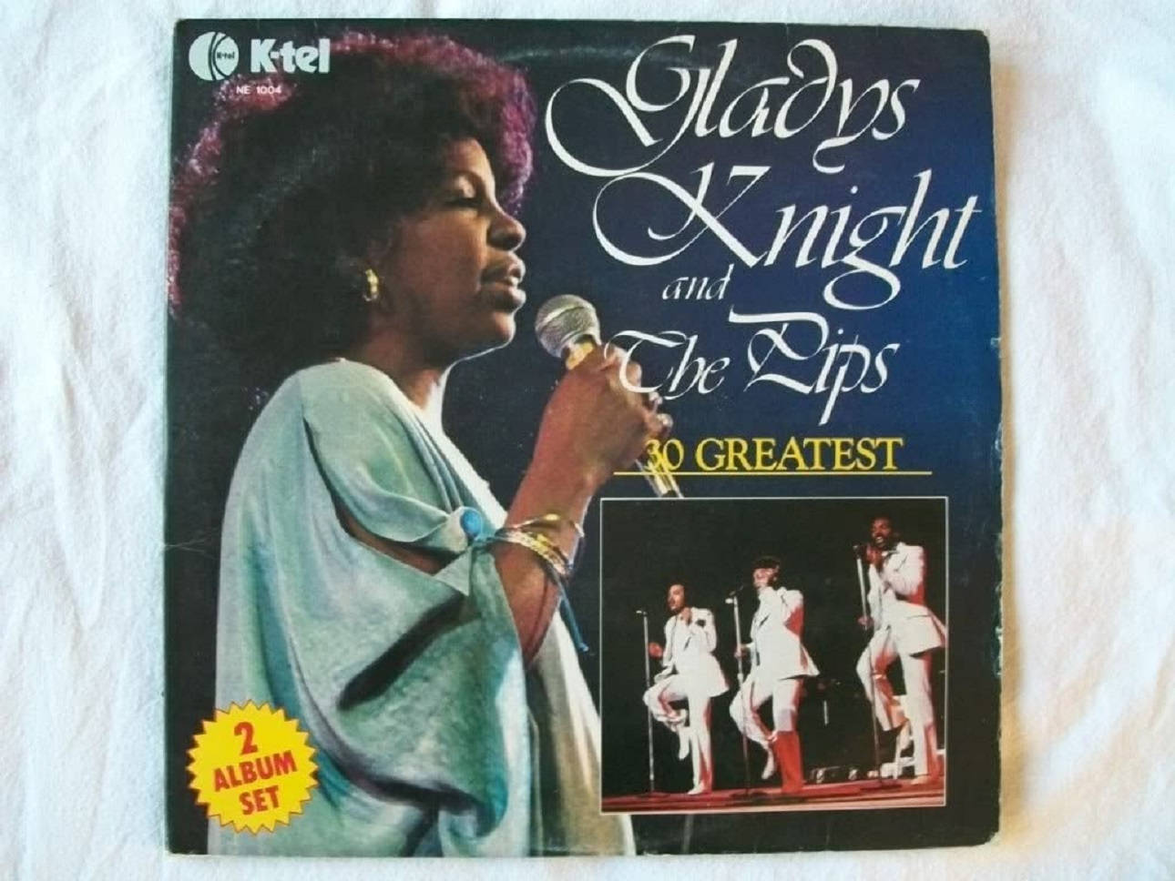 葛萊蒂絲·奈特與The Pips《30 Greatest Hits》專輯封面 壁紙 — Gladys Knight 身穿藍綠色長袍，在復古專輯封面上對著麥克風唱歌，下方是她穿著白色西裝的樂隊。