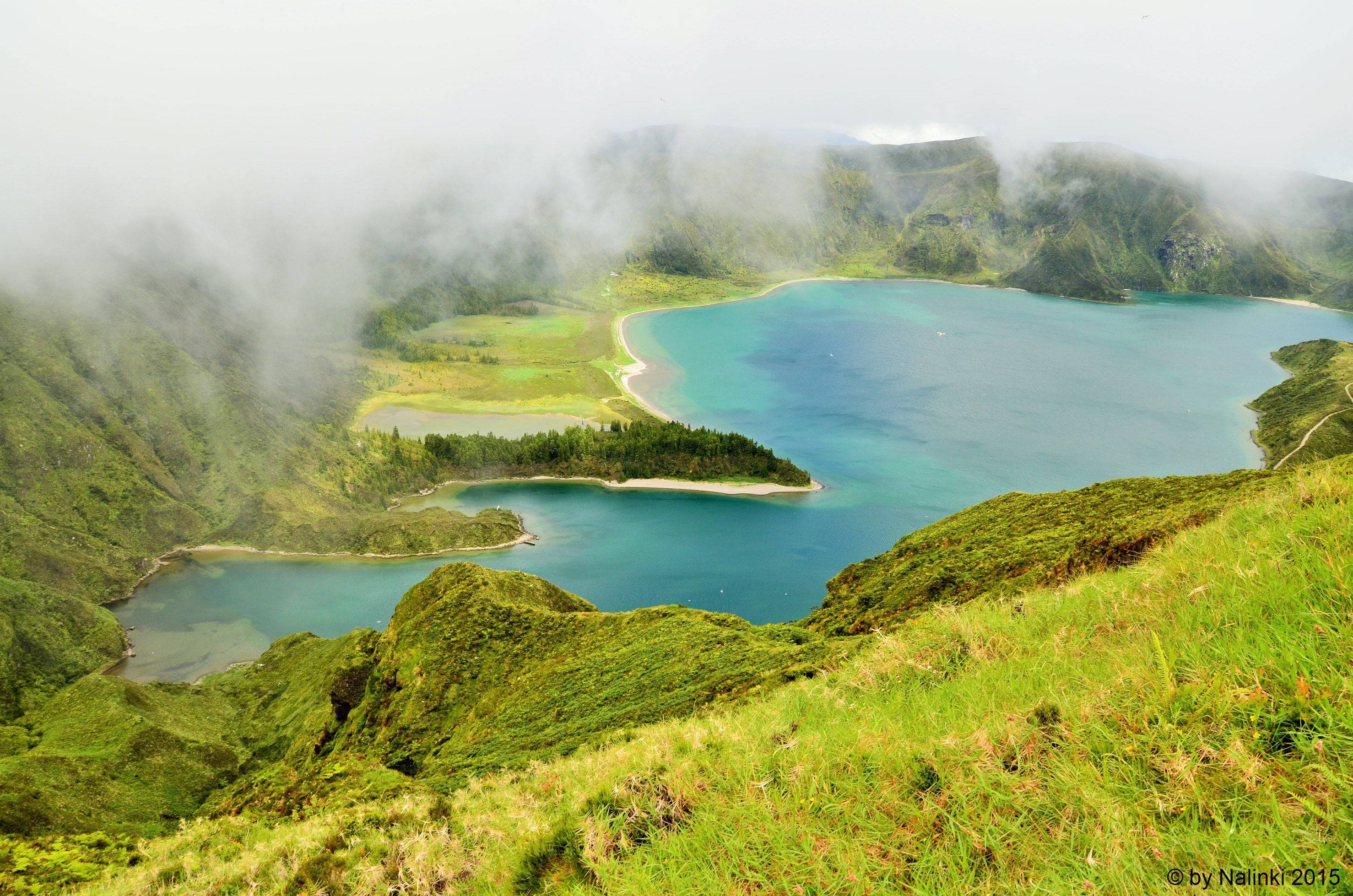葡萄牙聖米格尔島火湖 壁紙 — 一個霧氣瀰漫的火山口湖，湖水呈綠松石色，坐落在鬱鬱蔥蔥的綠色山丘之間，天空多雲。