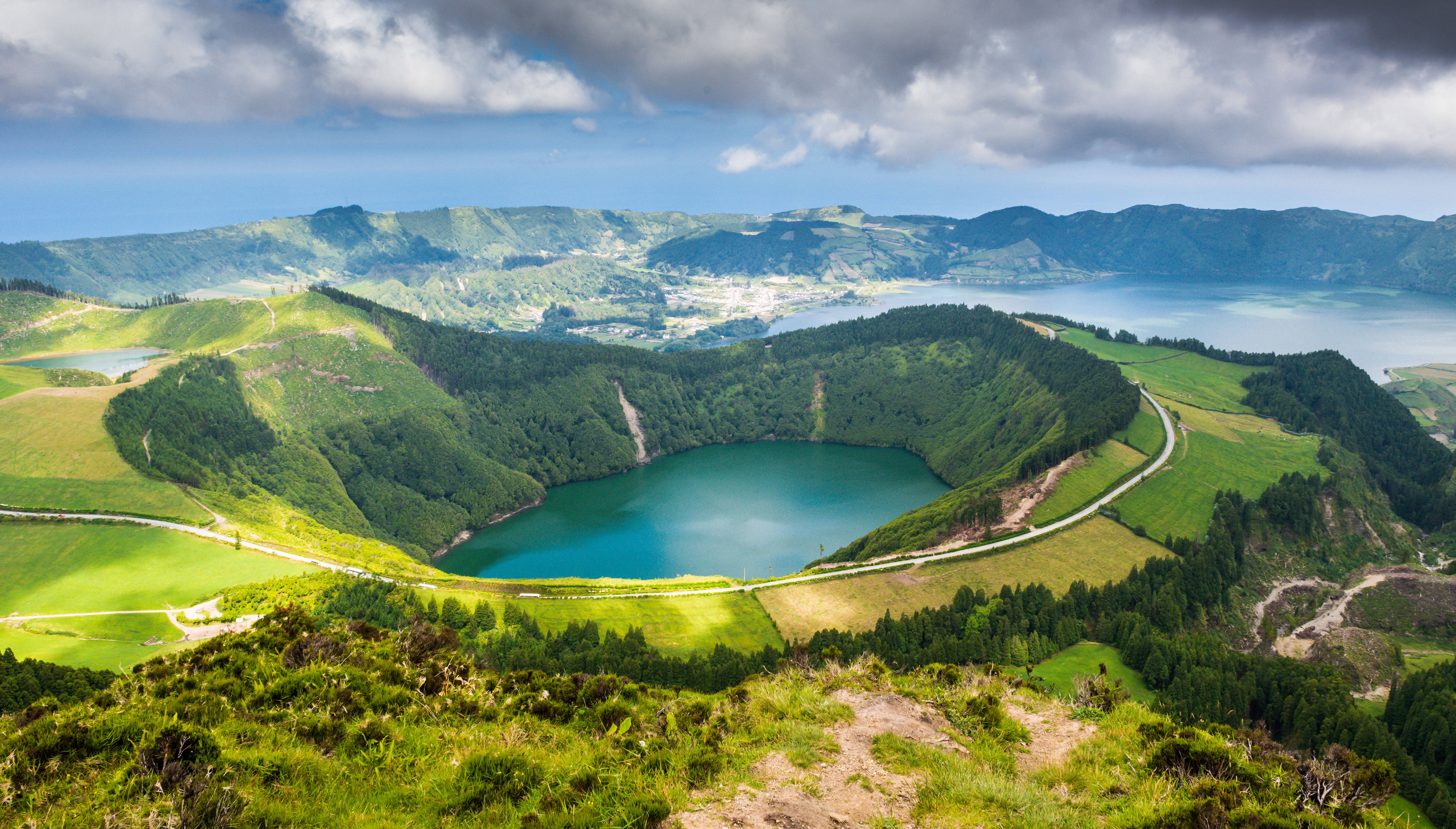 葡萄牙聖讓湖 壁紙 — 一個充滿活力的綠松色火山口湖，坐落在鬱鬱蔥蔥的綠色山丘中，上方是戲劇性的多雲天空。
