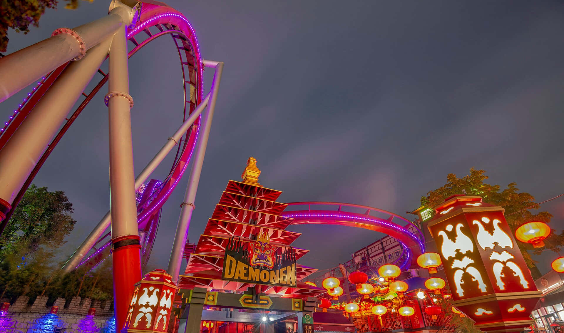 蒂沃利花園「惡魔」過山車夜景 壁紙 — 一個充滿活力的遊樂園夜景，特色是發光的紫色過山車拱起在一個裝飾著燈籠的紅色寶塔風格入口上方。