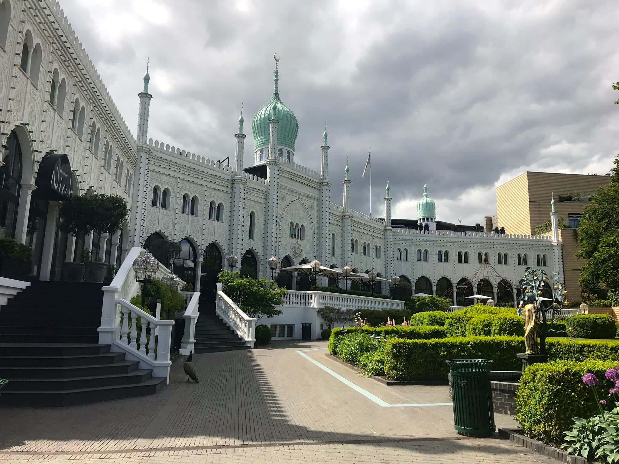 蒂沃利花園旁尼姆酒店的東側 壁紙 — 一座宏偉的白色鄂圖曼風格建築，擁有藍綠色圓頂和宣禮塔，矗立在多雲的天空下，兩側是修剪整齊的樹籬和石階梯。