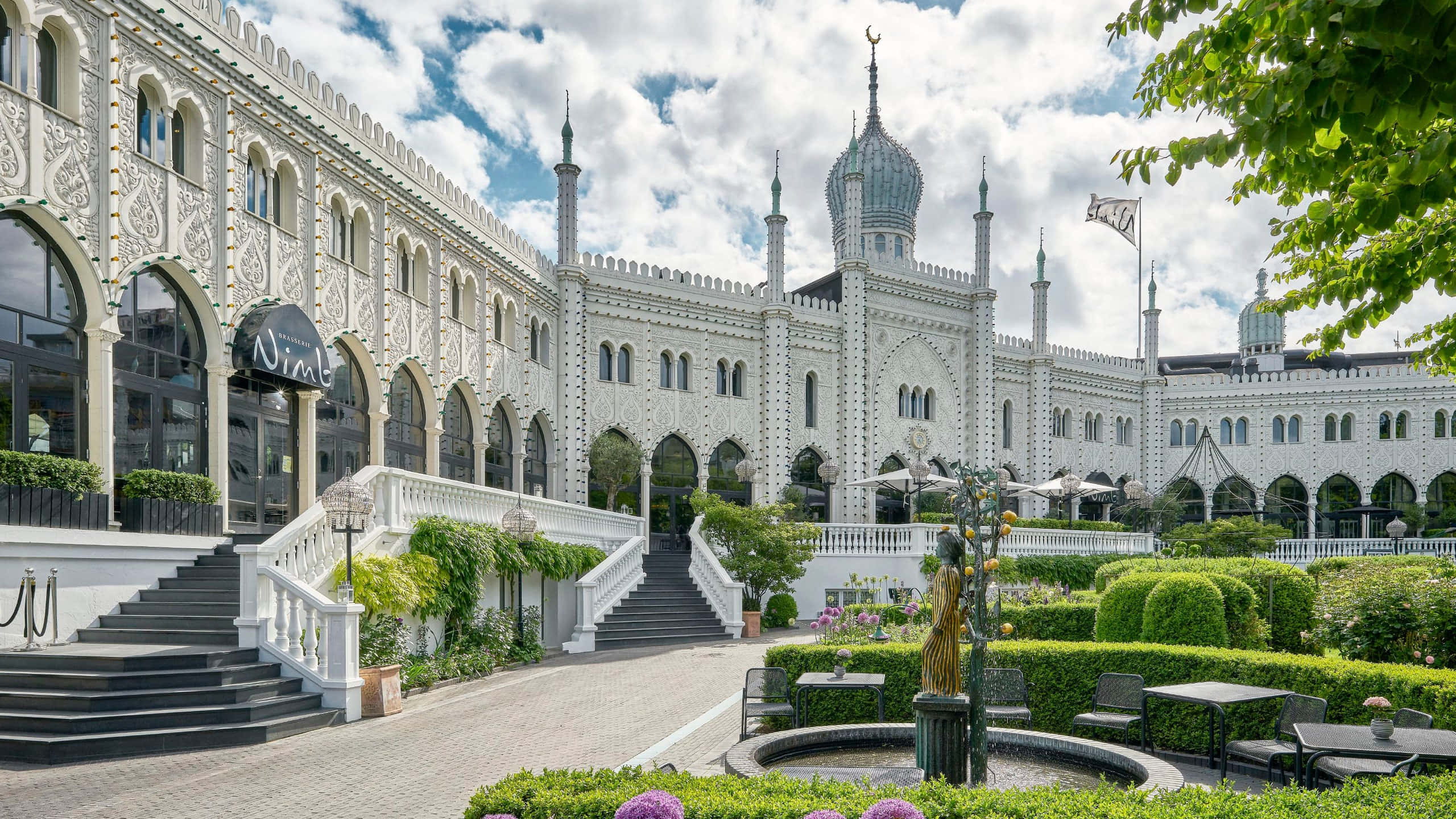 蒂沃利花園旁的尼姆酒店階梯 壁紙 — 宏偉的白色鄂圖曼風格建築，擁有華麗的拱門和圓頂，天空部分多雲，周圍是修剪整齊的花園和戶外座位區。
