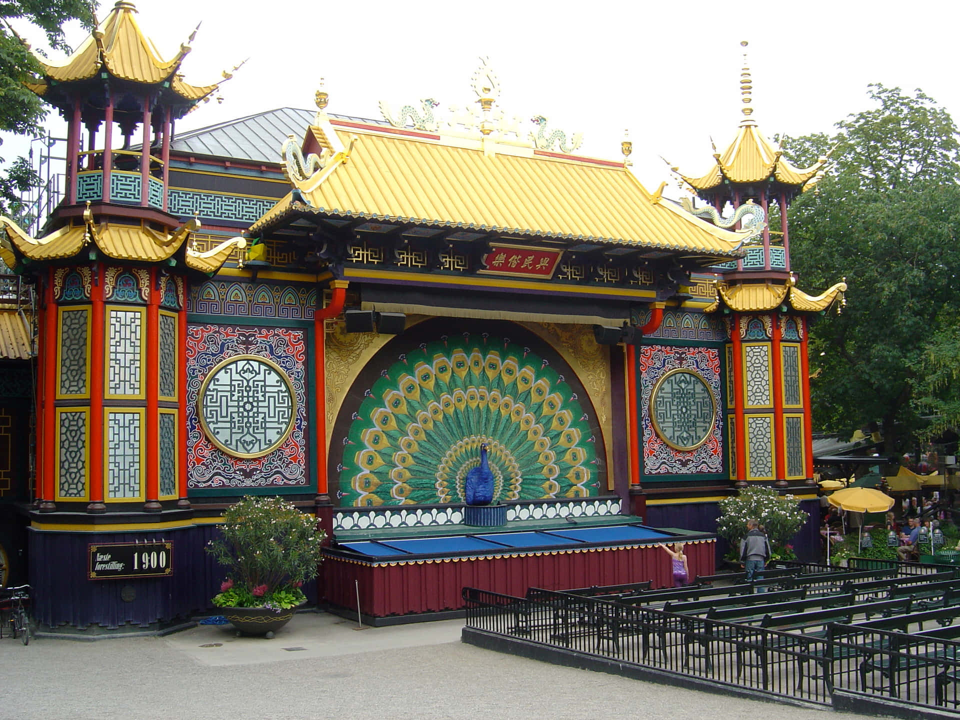 蒂沃利花園木偶劇院 壁紙 — 一座充滿活力的中式亭閣，金色屋頂和大型孔雀壁畫，矗立在公園中，周圍有綠樹和遊客。