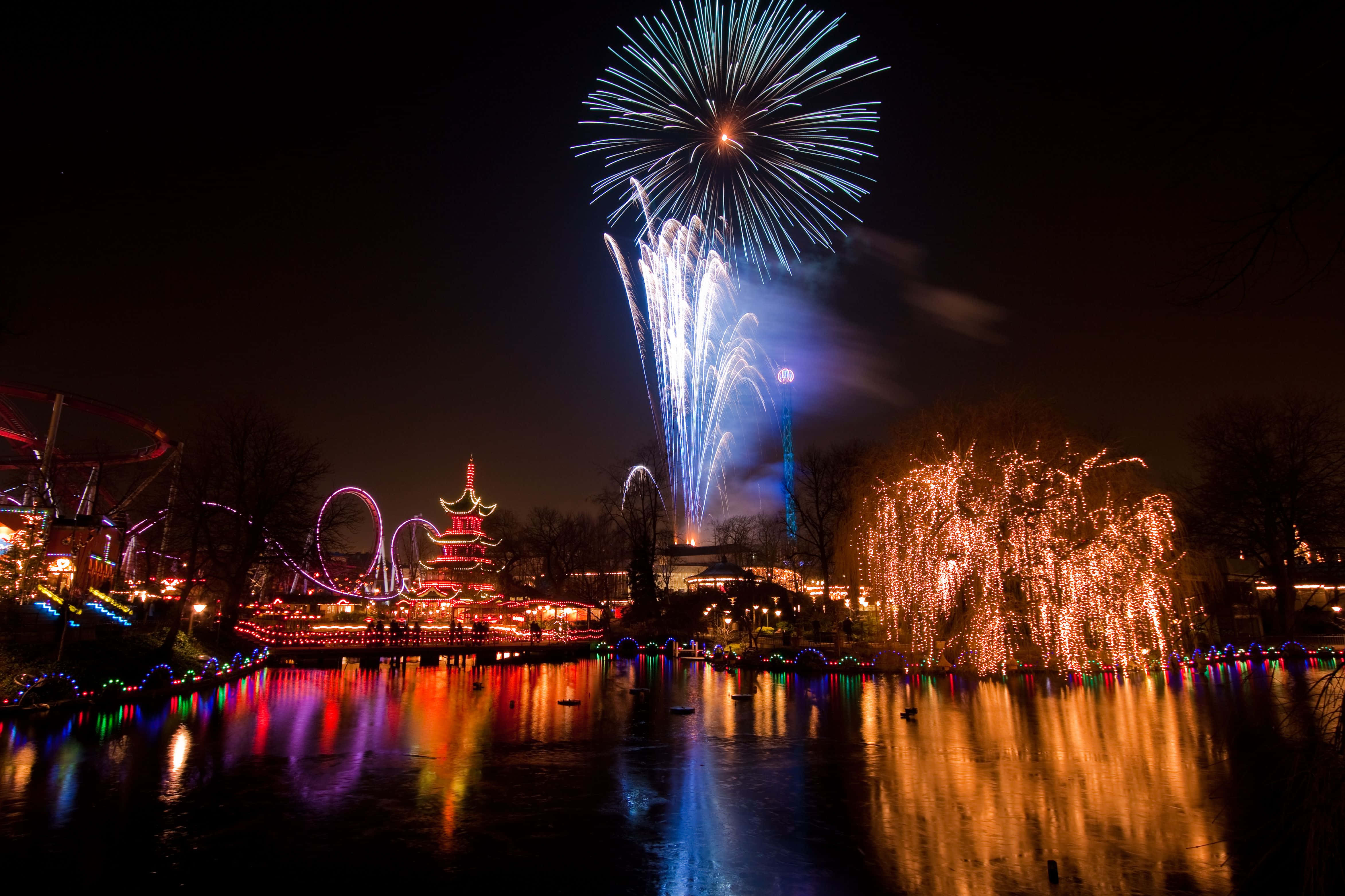 蒂沃利花園煙火與城市燈光 壁紙 — 色彩繽紛的煙火在夜間遊樂園上空綻放，倒映在平靜的水面上。
