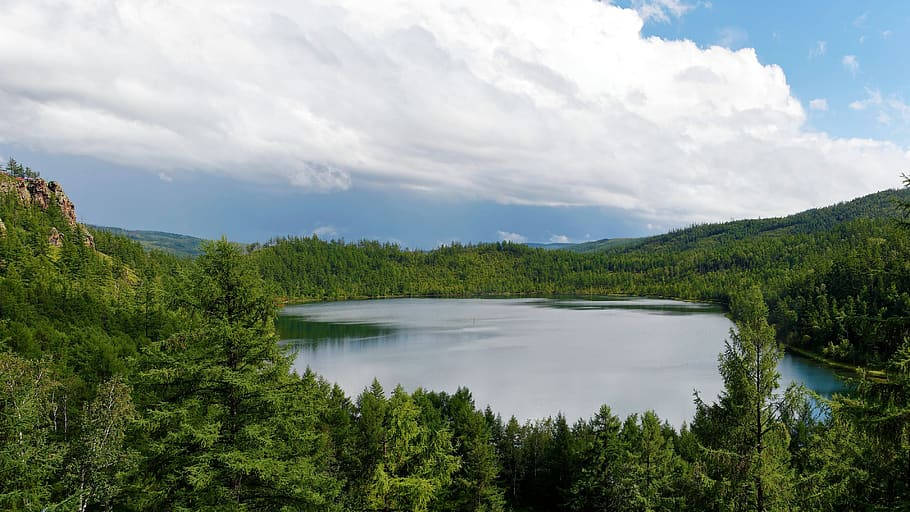 蒙古地區的湖景 壁紙 — 一個寧靜的湖泊坐落在茂密的綠色森林中，天空部分多雲，遠處有山丘。