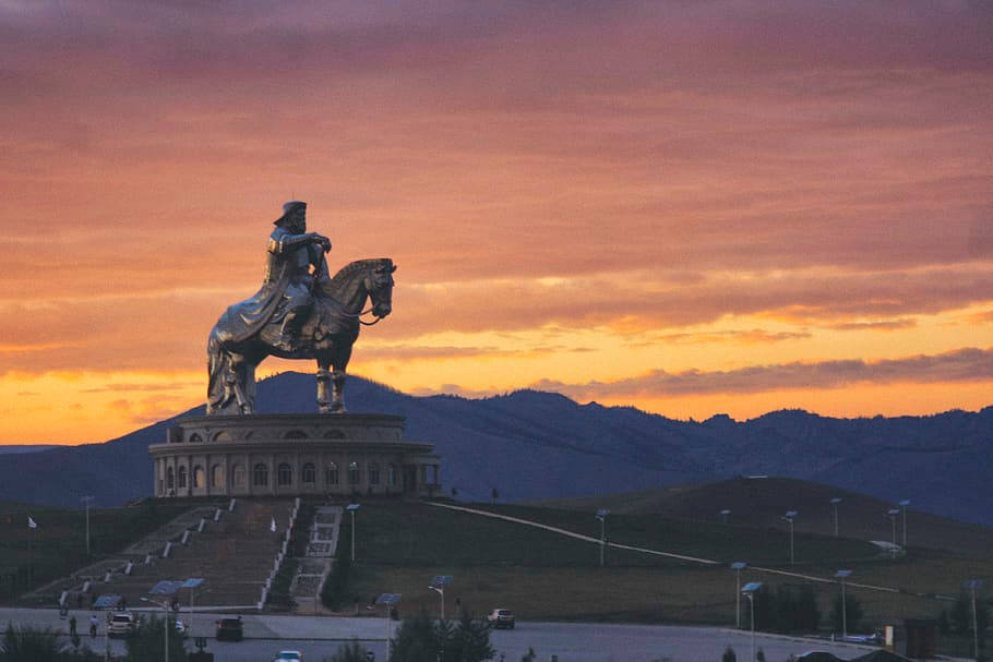 蒙古成吉思汗雕像迎夕陽 壁紙 — 一座雄偉的青銅騎馬雕像矗立在宏偉的基座上，背景是熾熱的日落天空和遠處連綿起伏的山巒。