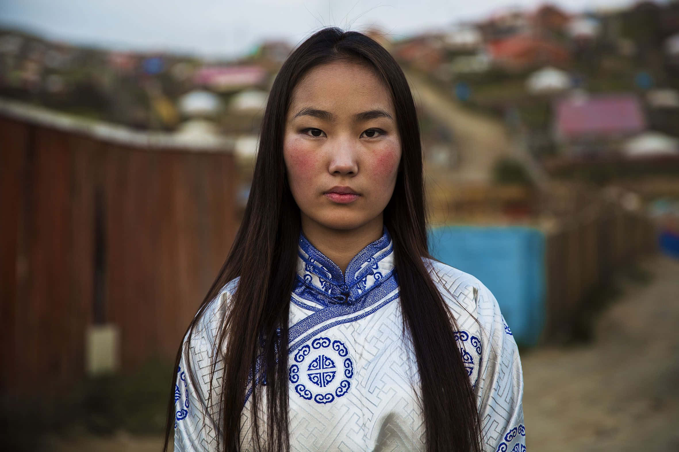 蒙古族美女文化風情圖 壁紙 — 一位身穿傳統藍白刺繡服飾的年輕女子，在模糊的山坡村莊背景下莊嚴地站立。