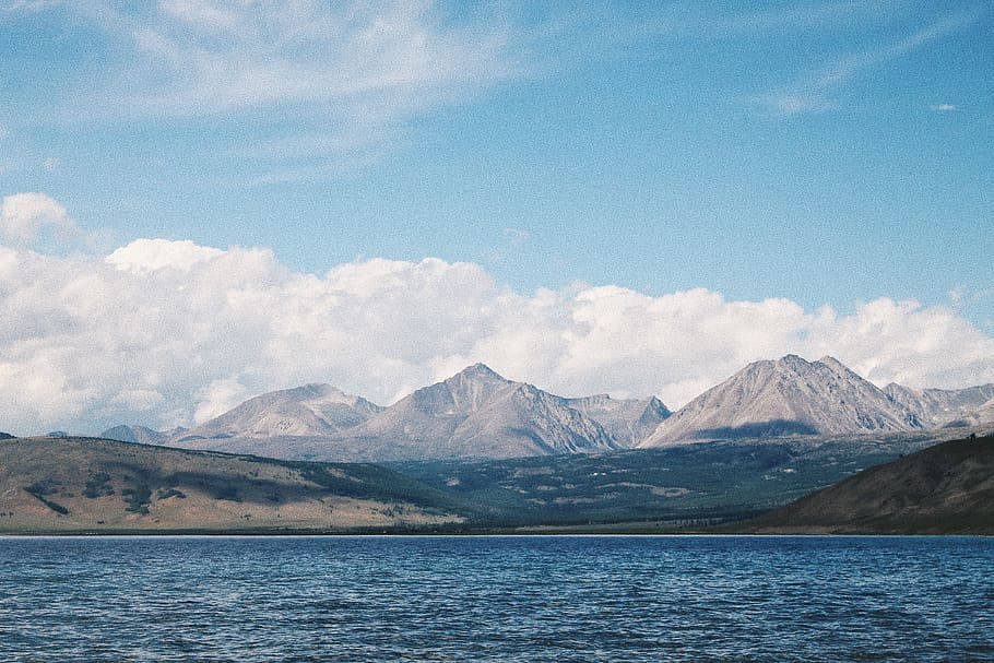 蒙古湖上雲影 壁紙 — 一個寧靜的湖泊倒映著廣闊的天空，在明亮的日光下，崎嶇的山峰上方飄著蓬鬆的雲朵。