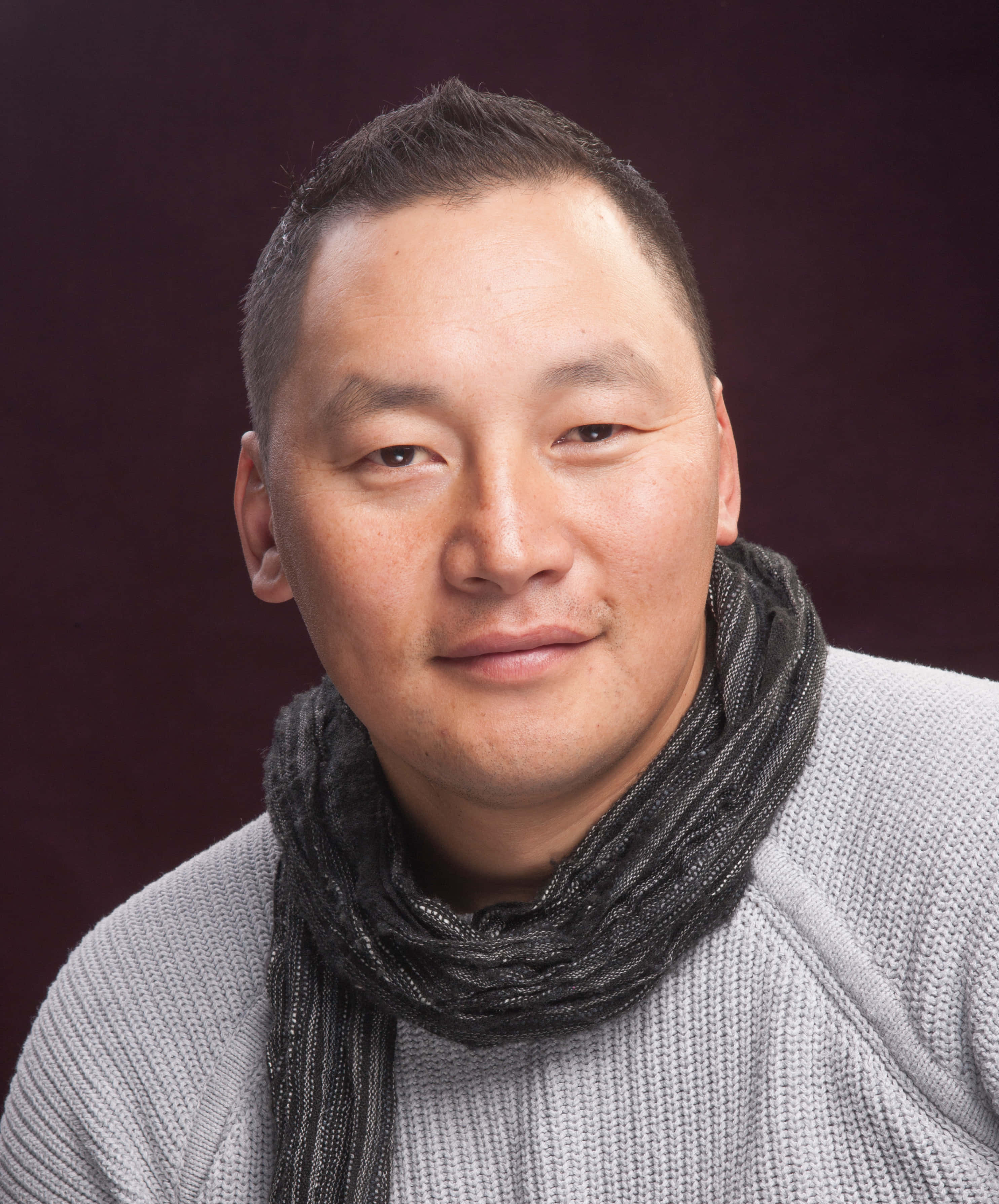 蒙古演員Baljinnyamyn Amarsaikhan肖像 壁紙 — 一位短黑髮男士穿著灰色毛衣和深色圍巾，背景是深酒紅色的攝影棚。