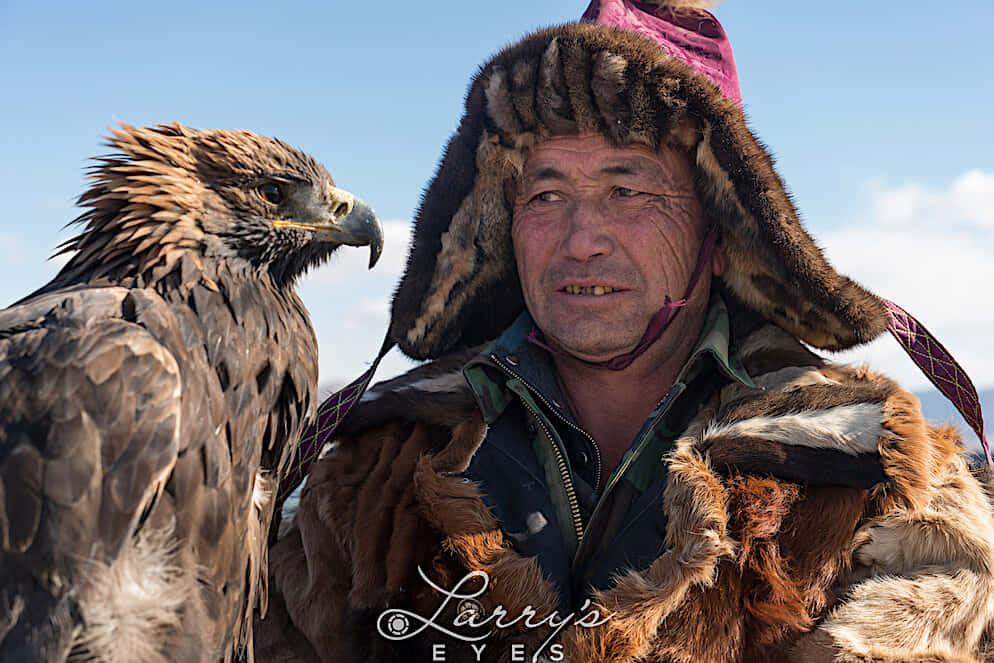 蒙古鷹獵者與野生動物 壁紙 — 一位飽經風霜的鷹獵人，戴著皮帽、穿著大衣，站在他雄偉的金雕旁邊，背景是清澈的藍天。