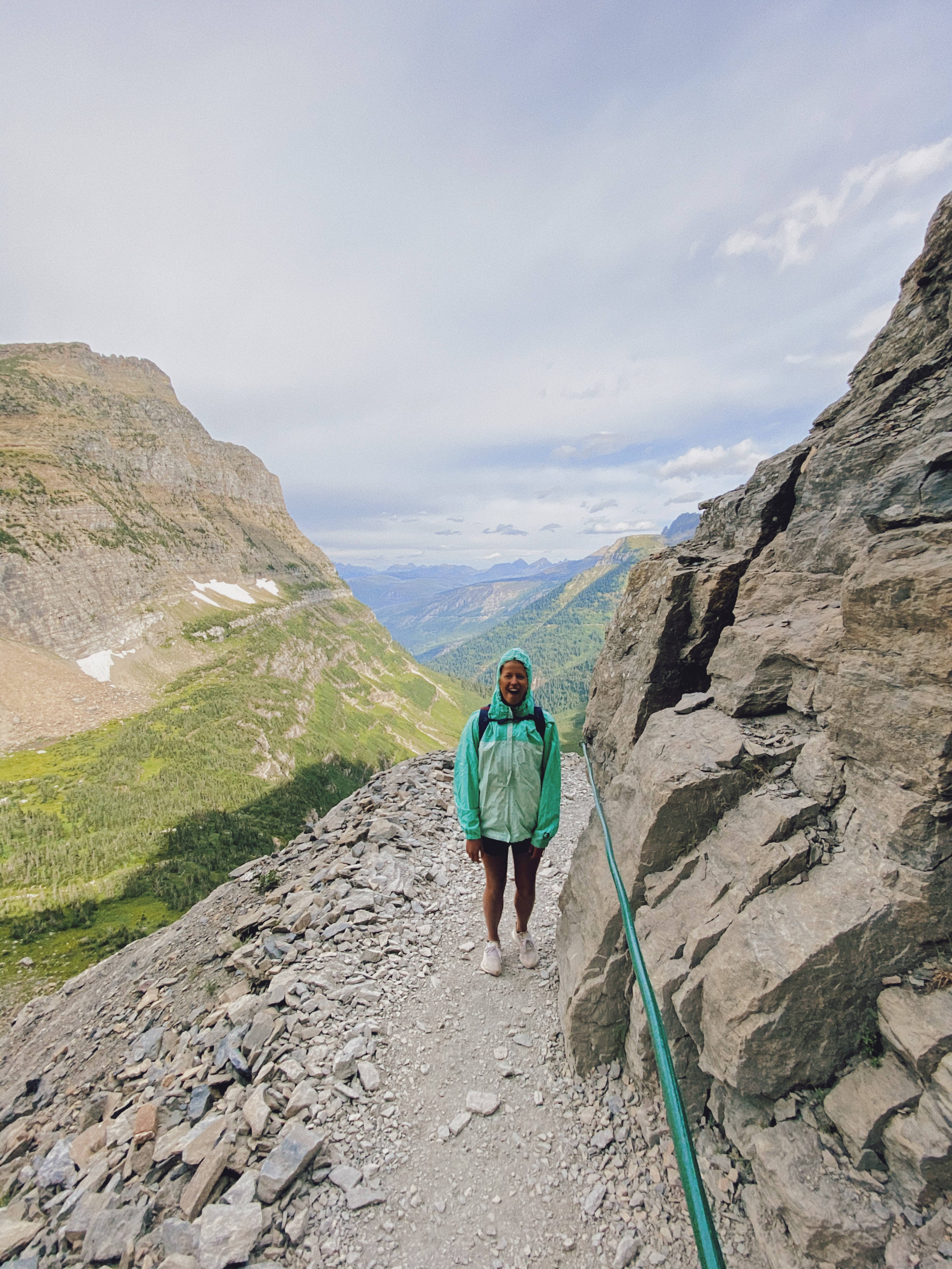 蒙大拿州女登山者 iPhone 壁紙 — 一位穿著青綠色夾克的徒步旅行者在多岩石的山路上微笑，周圍有陡峭的懸崖和遠處的綠色山谷，天空多雲。