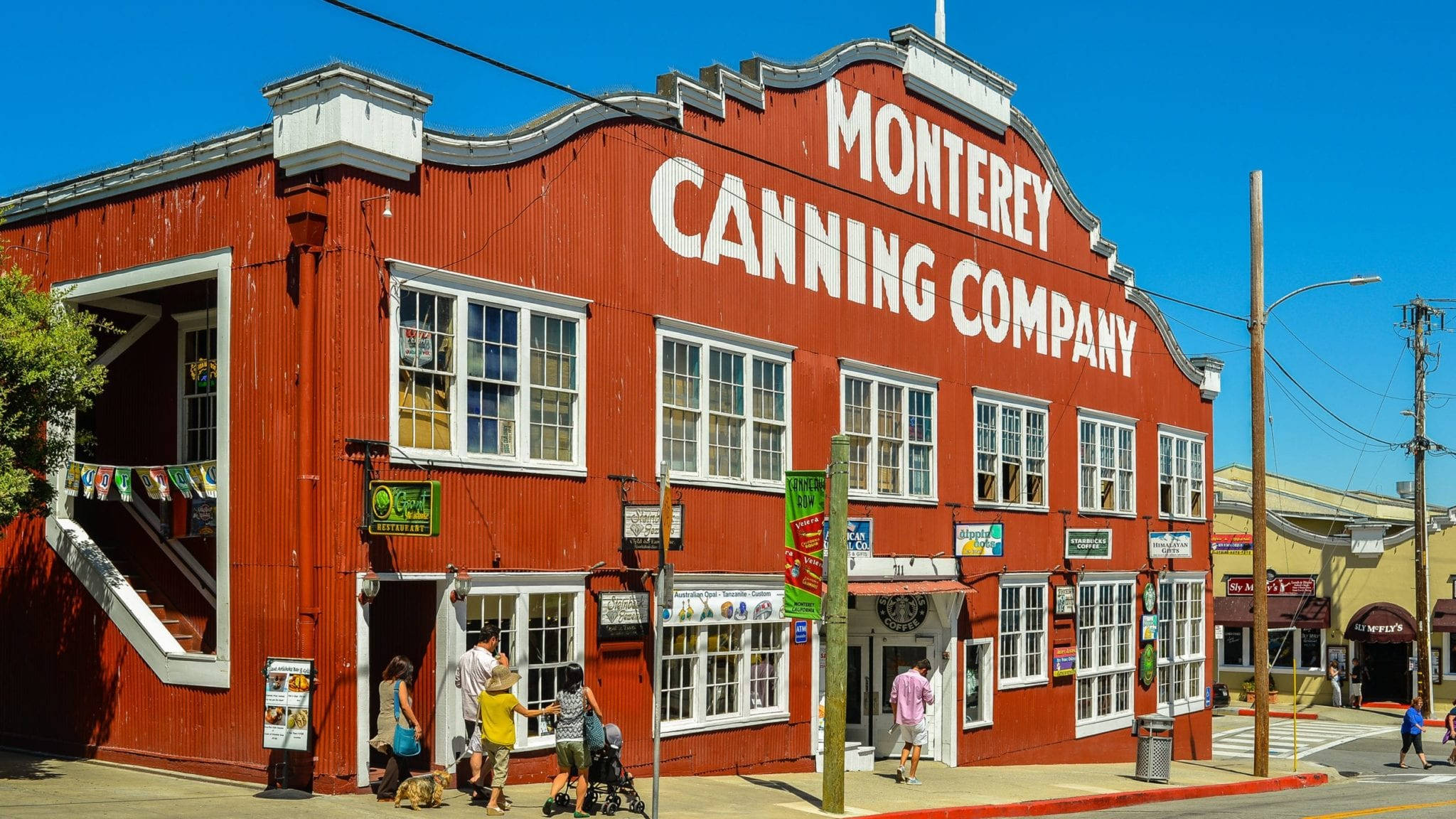 蒙特雷罐頭公司大樓 壁紙 — 一座鮮紅色的歷史建築 Monterey Canning Company，帶有白色飾邊，人們在晴朗的藍天下走在行人路上。