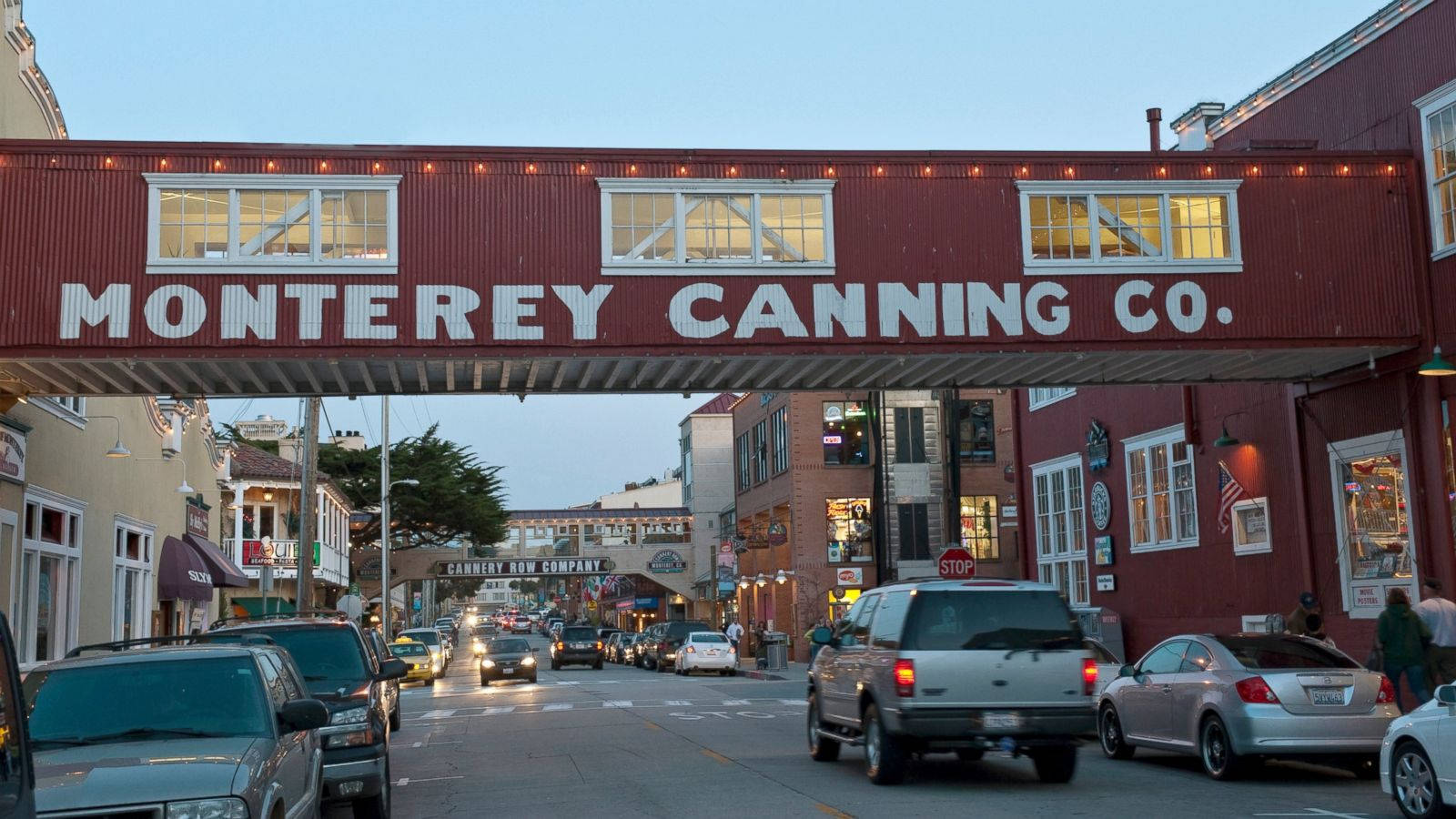 蒙特雷罐頭廠在坎農行街 壁紙 — 一座紅色的 Monterey Canning Co. 行人天橋橫跨一條繁忙的街道，街道上有汽車和商店門面，正值黃昏時分。