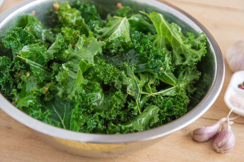蒜香羽衣甘藍食譜 壁紙 — 新鮮的羽衣甘藍葉在不銹鋼碗中閃閃發光，旁邊是蒜瓣，放在木質檯面上。
