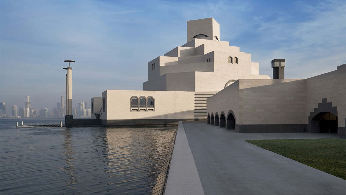蔚藍天空下的壯麗伊斯蘭藝術博物館 壁紙 — 現代白色幾何博物館建築，帶有拱形窗戶，矗立在海濱，反射天空和城市天際線的背景。