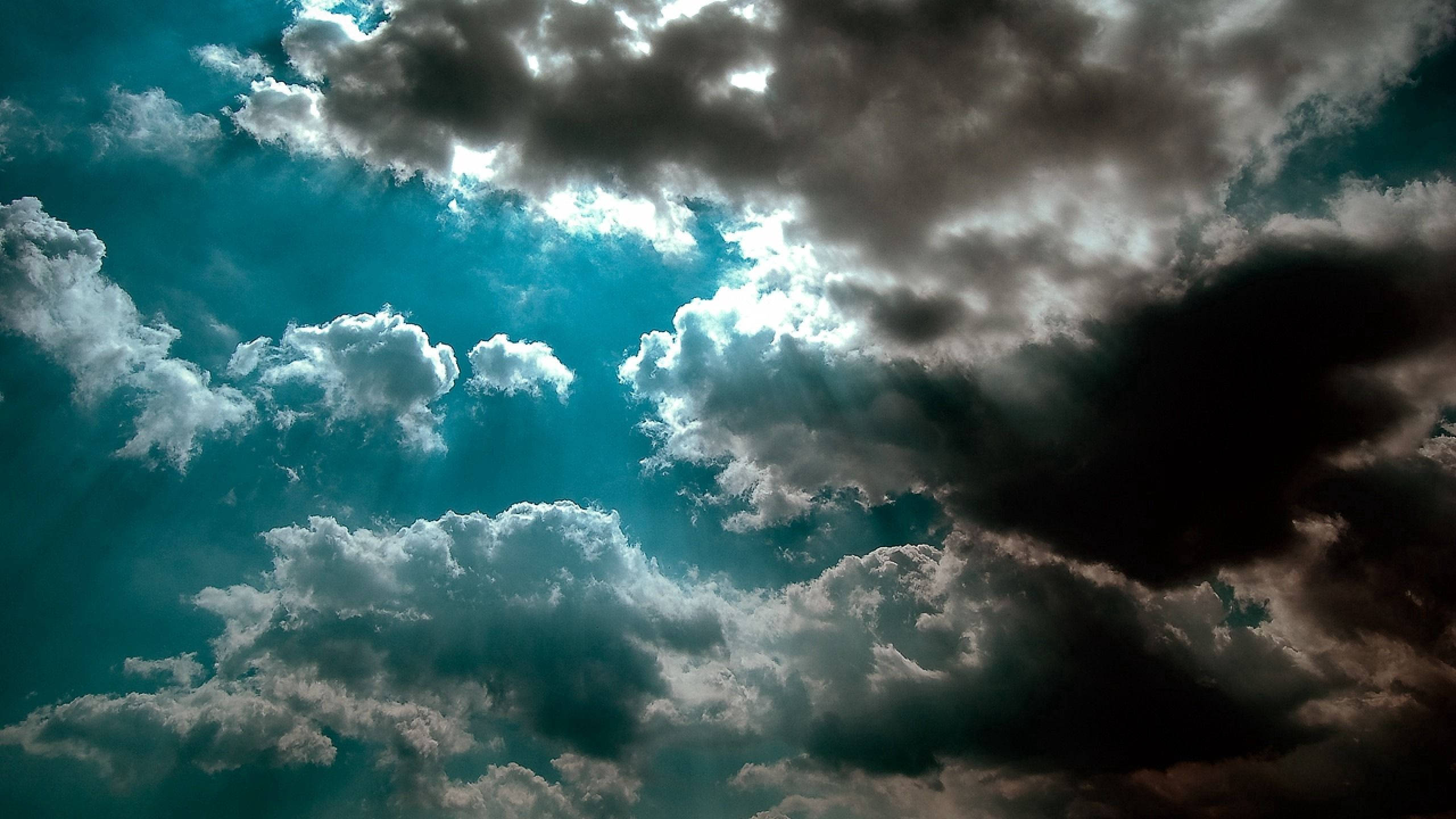 蔚藍天空中的烏雲 壁紙 — 戲劇性的暴風雲層，陽光穿透青綠色的天空，營造出陰鬱而強大的氛圍。