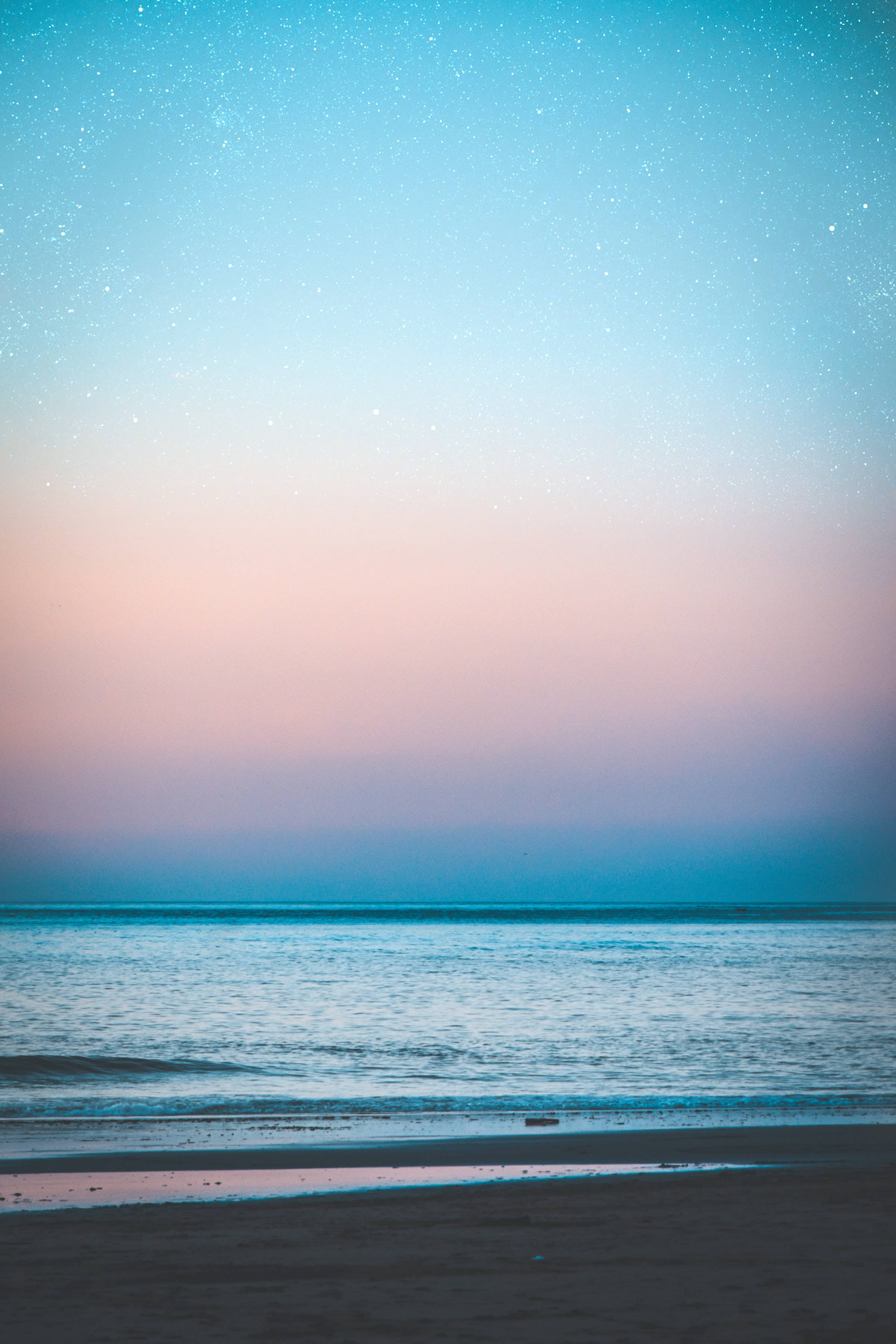 蔚藍海洋與繁星夜空 壁紙 — 暮色中寧靜的海灘，星空從粉紅過渡到藍色，映照在平靜的水面上。