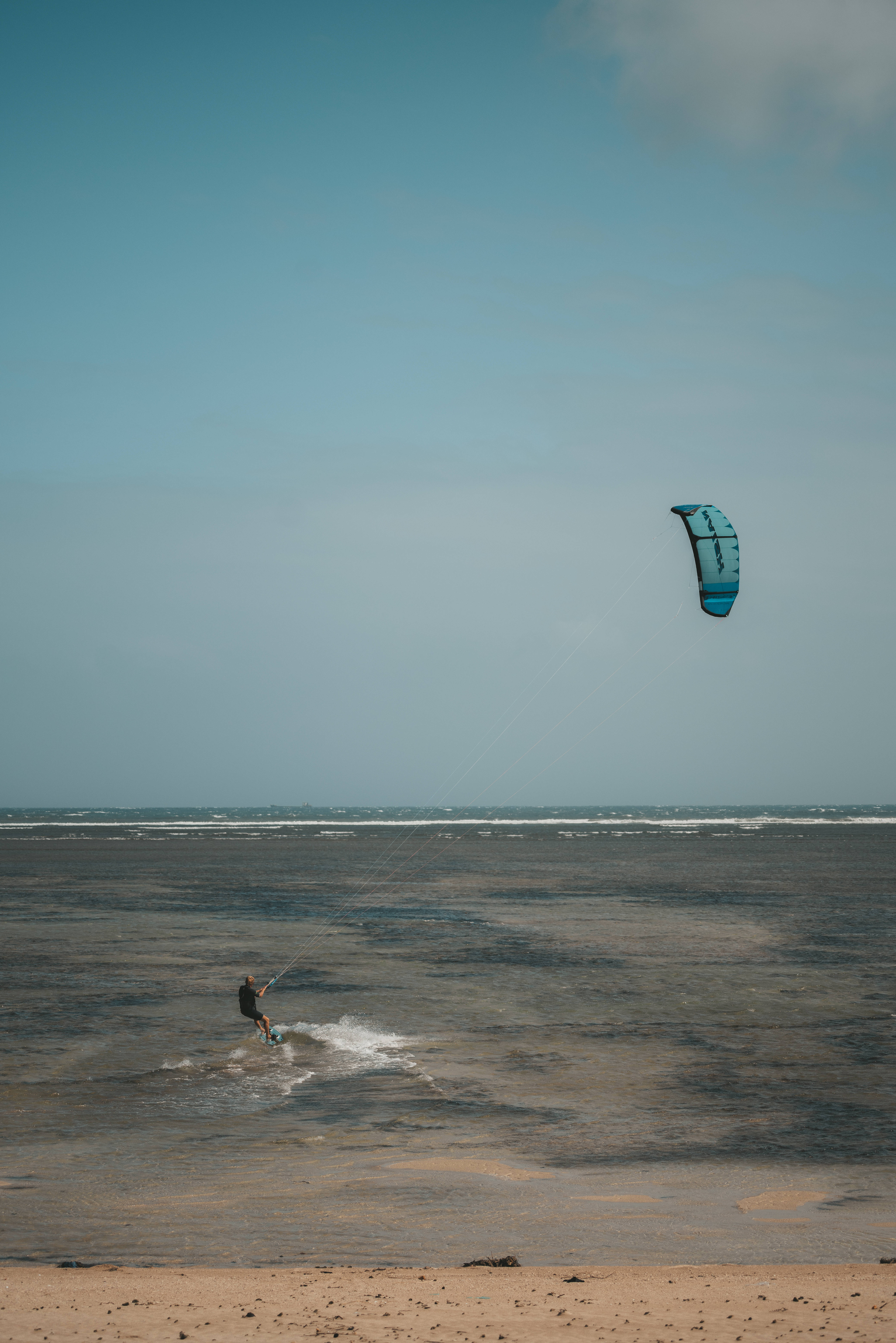 蔚藍海洋風帆衝浪 壁紙 — 一名風箏衝浪者在廣闊的藍天下，於淺水沿岸水域滑行，頭頂上有一隻充滿活力的藍色風箏翱翔。