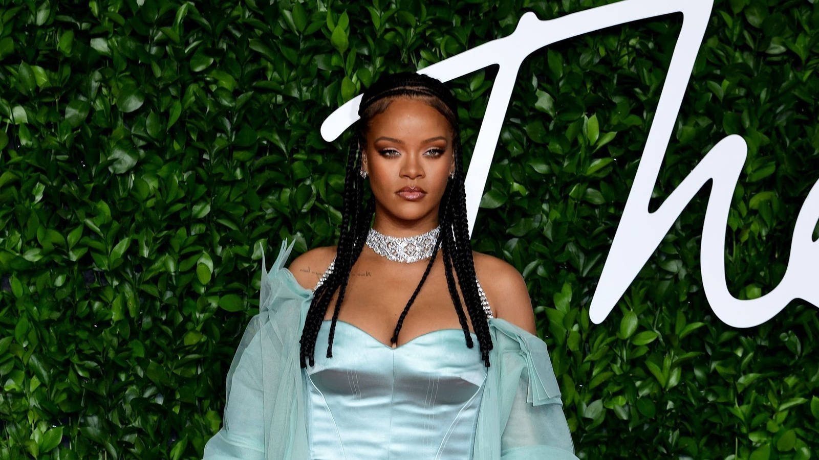Rihanna 身穿藍色緞面長袍，辮子和頸鍊，背景是綠色樹籬。