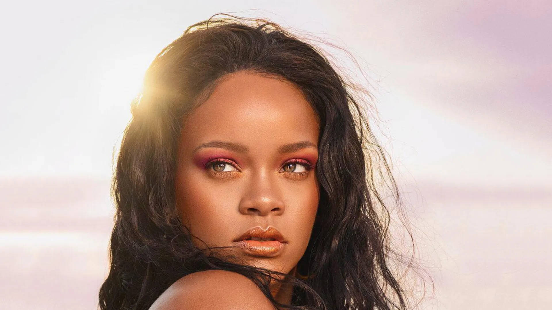 Rihanna 塗上粉紅色眼影，頭髮在陽光下閃耀，背景是粉彩天空。