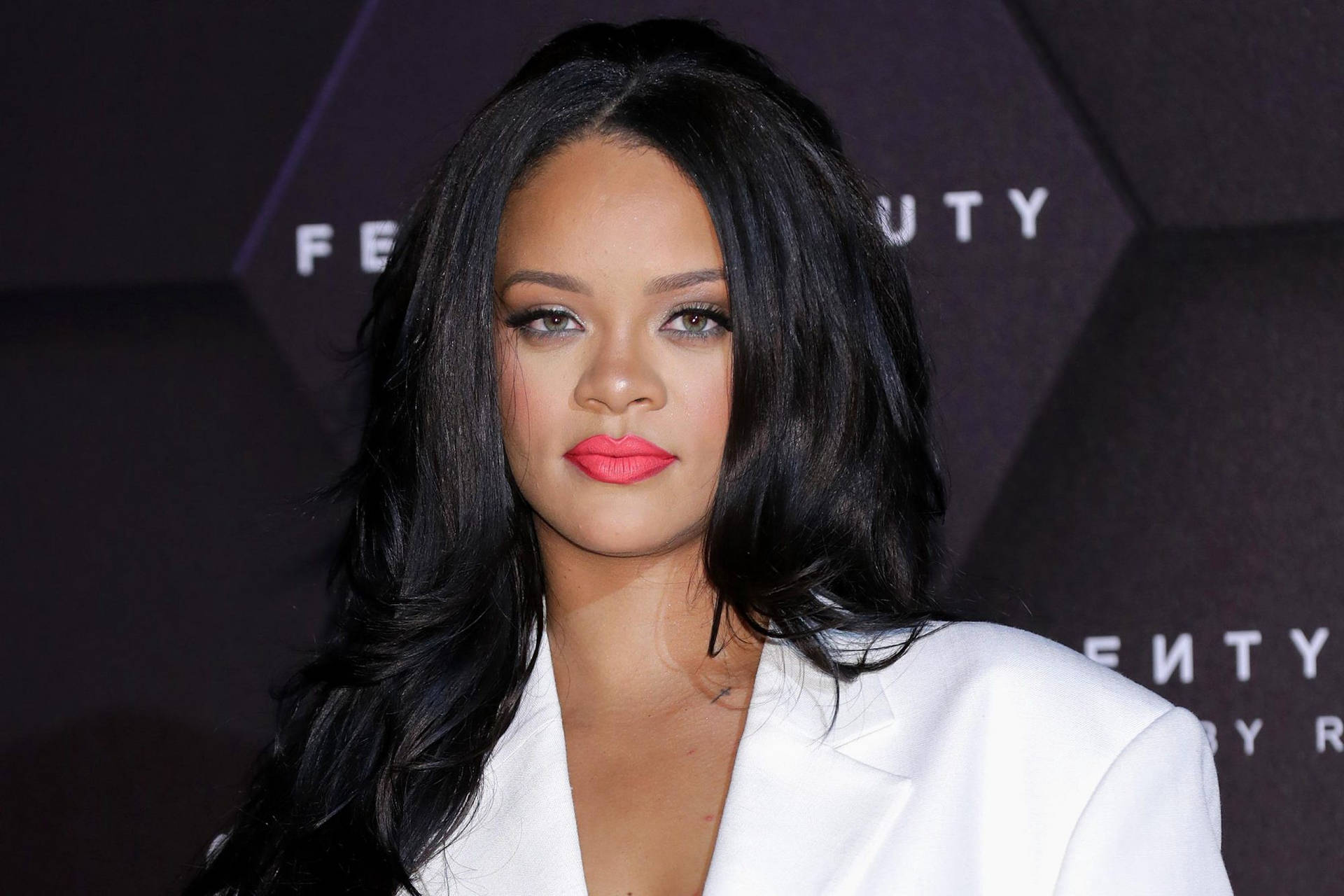 Rihanna 在 Fenty Beauty 活動中穿著白色西裝外套，擁有鮮豔的粉紅嘴唇和黑髮。