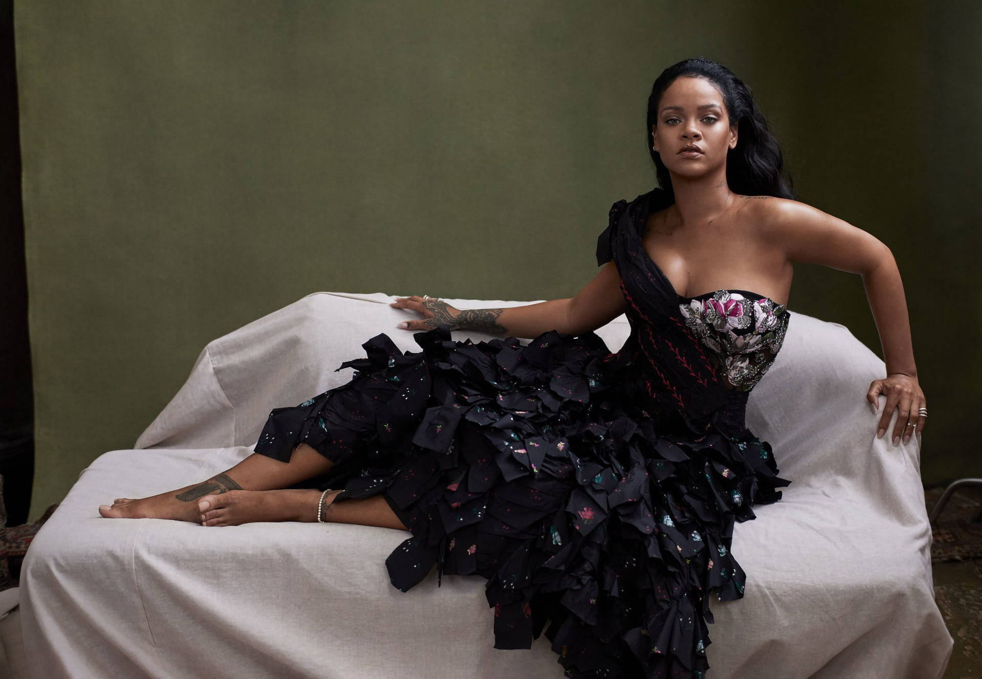 Rihanna 身穿黑色荷葉邊禮服，躺在一張白色垂簾沙發上。