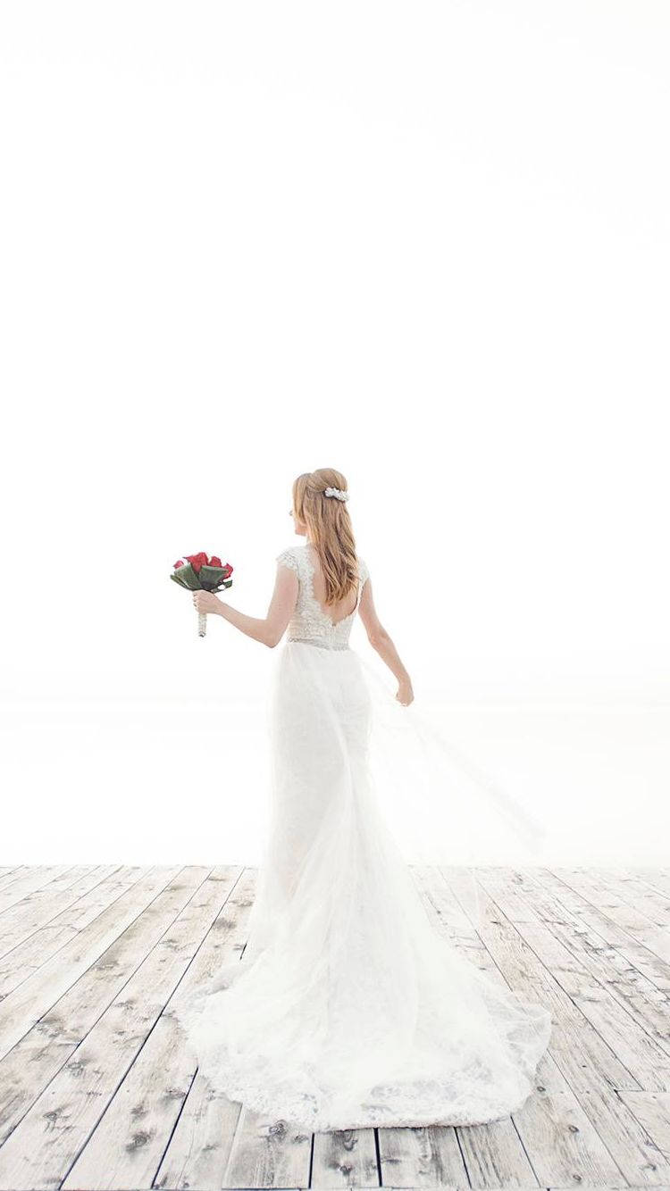 蕾絲短袖婚紗 壁紙 — 一位身穿飄逸白色婚紗的新娘，在風化的木甲板上手持一束紅玫瑰，背對著明亮的白色背景走開。