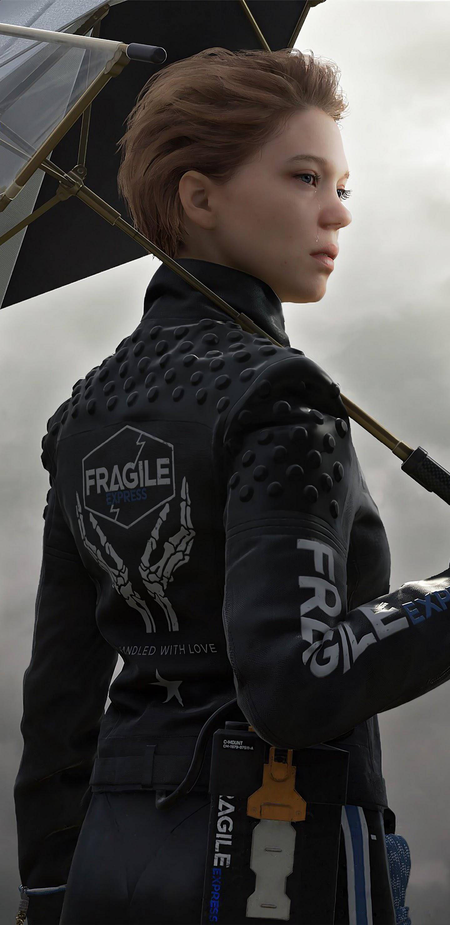 蕾雅·賽杜飾演角色「Fragile」 壁紙 — 一名身穿黑色裝甲外套、帶有'FRAGILE'字樣的女性，在灰色天空下撐著雨傘。