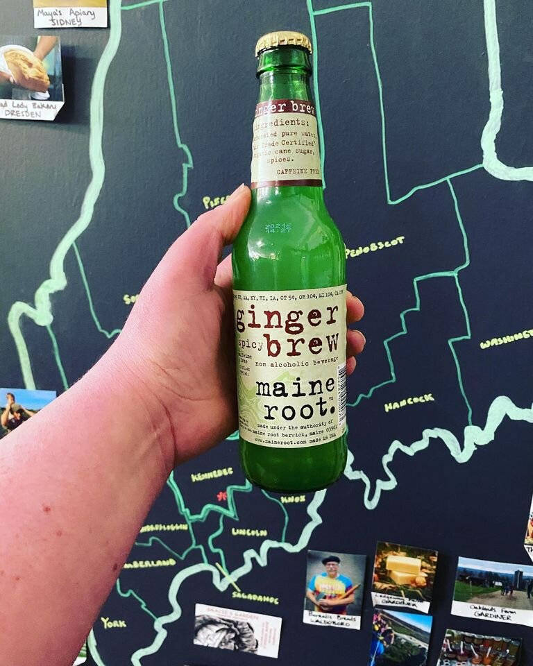 薑味根汁馬薩諸塞州飲品 背景為馬薩諸塞州地圖 壁紙 — 一隻手拿著一瓶綠色薑汁汽水，對著一面裝飾著旅行照片的深色地圖牆。